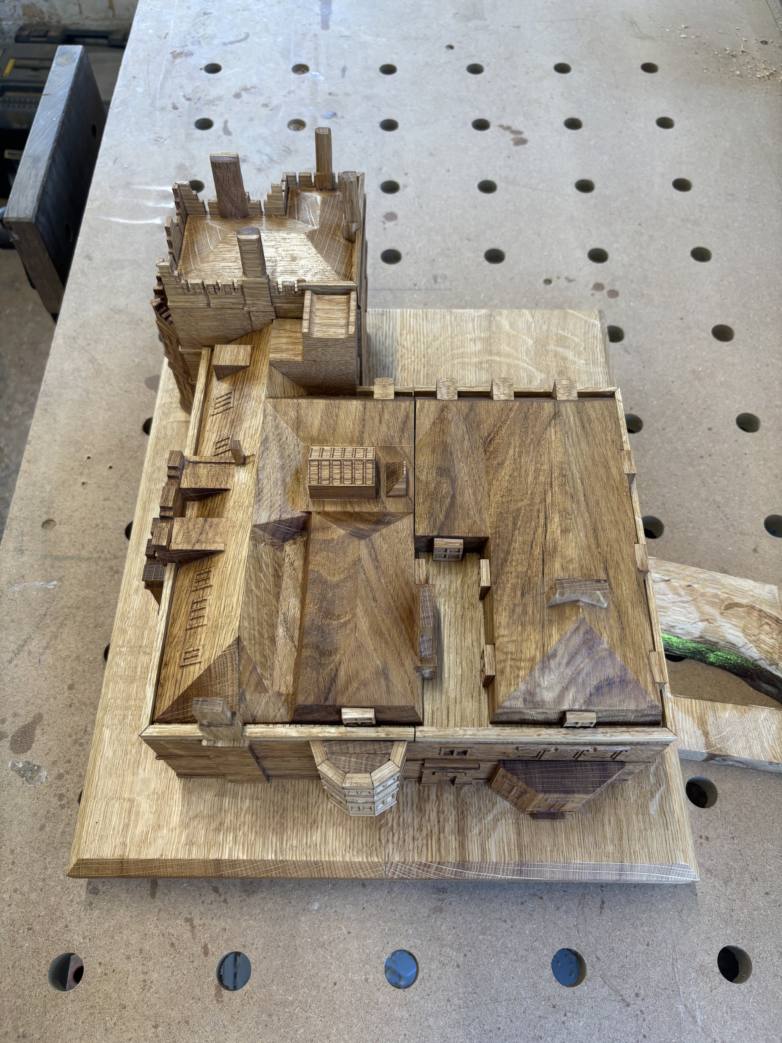 Wooden architectural model of a multi-story building with various rooms and features, placed on a workbench with a pegboard surface.