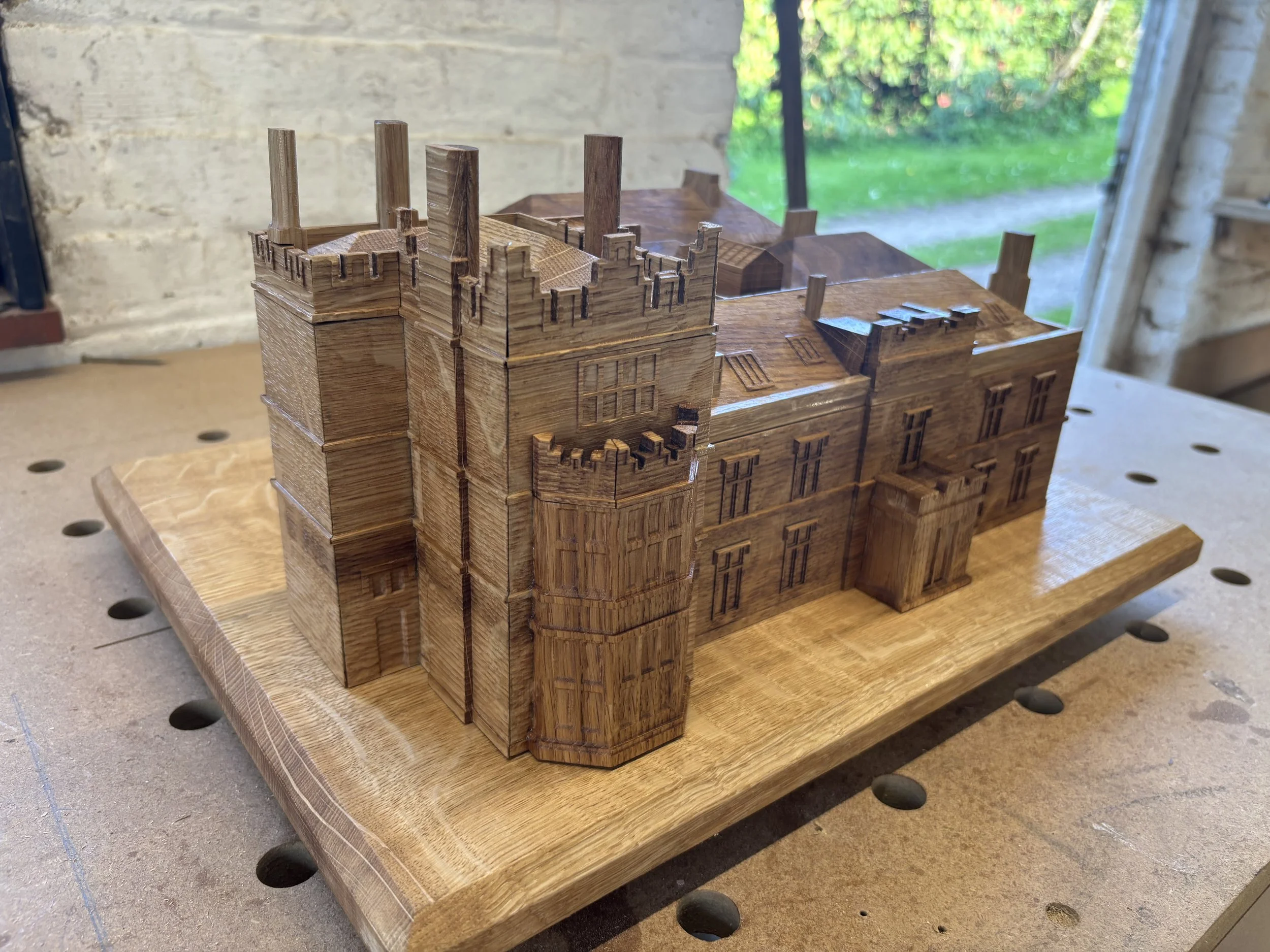 A detailed wooden model of a castle or fortress on a wooden base, placed on a workbench in a workshop with a window showing greenery outside.