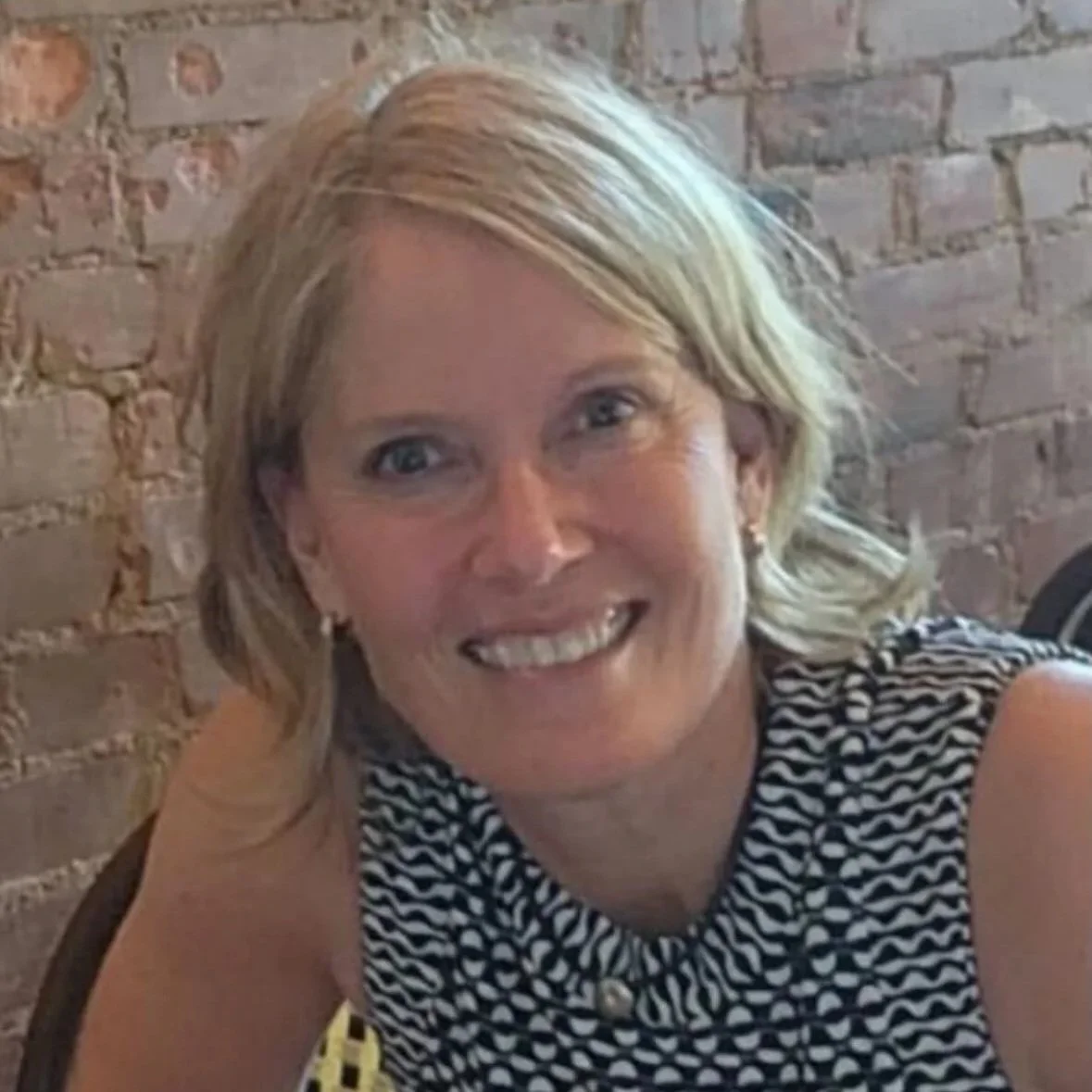 A smiling woman with blonde hair and blue eyes, wearing a black and white patterned sleeveless top, sitting in front of a brick wall.
