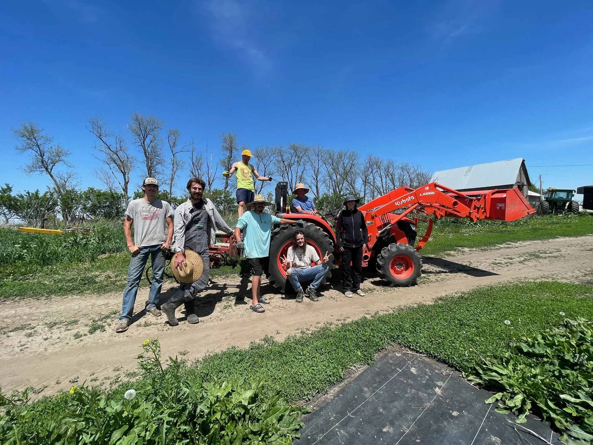 Group of seven people standing and sitting next to a red tractor on a farm, with green plants and a barn in the background, under a clear blue sky.