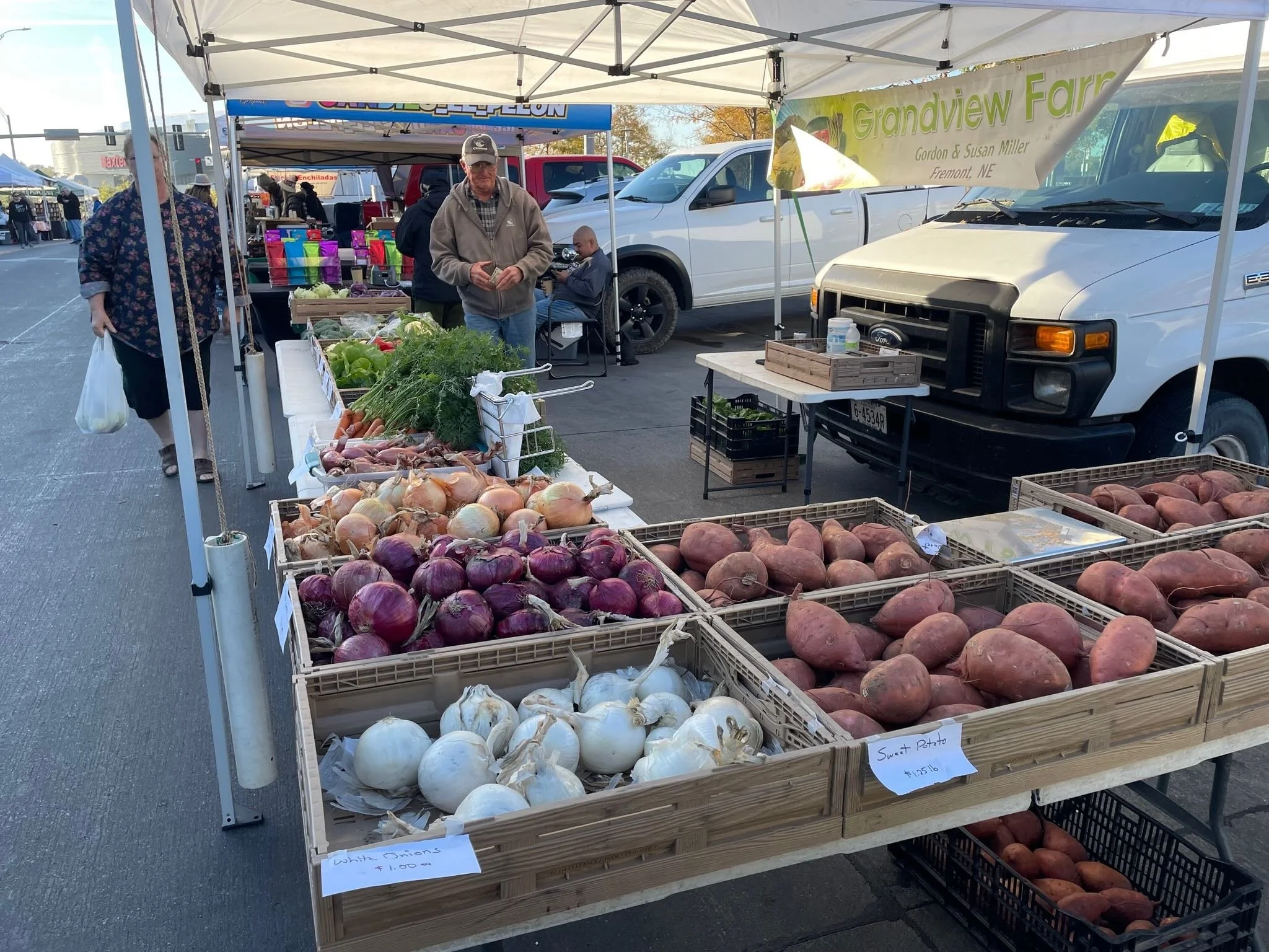 Outdoor farmers market stall displaying various fresh vegetables including white onions, red onions, sweet potatoes, and yams, with people shopping.