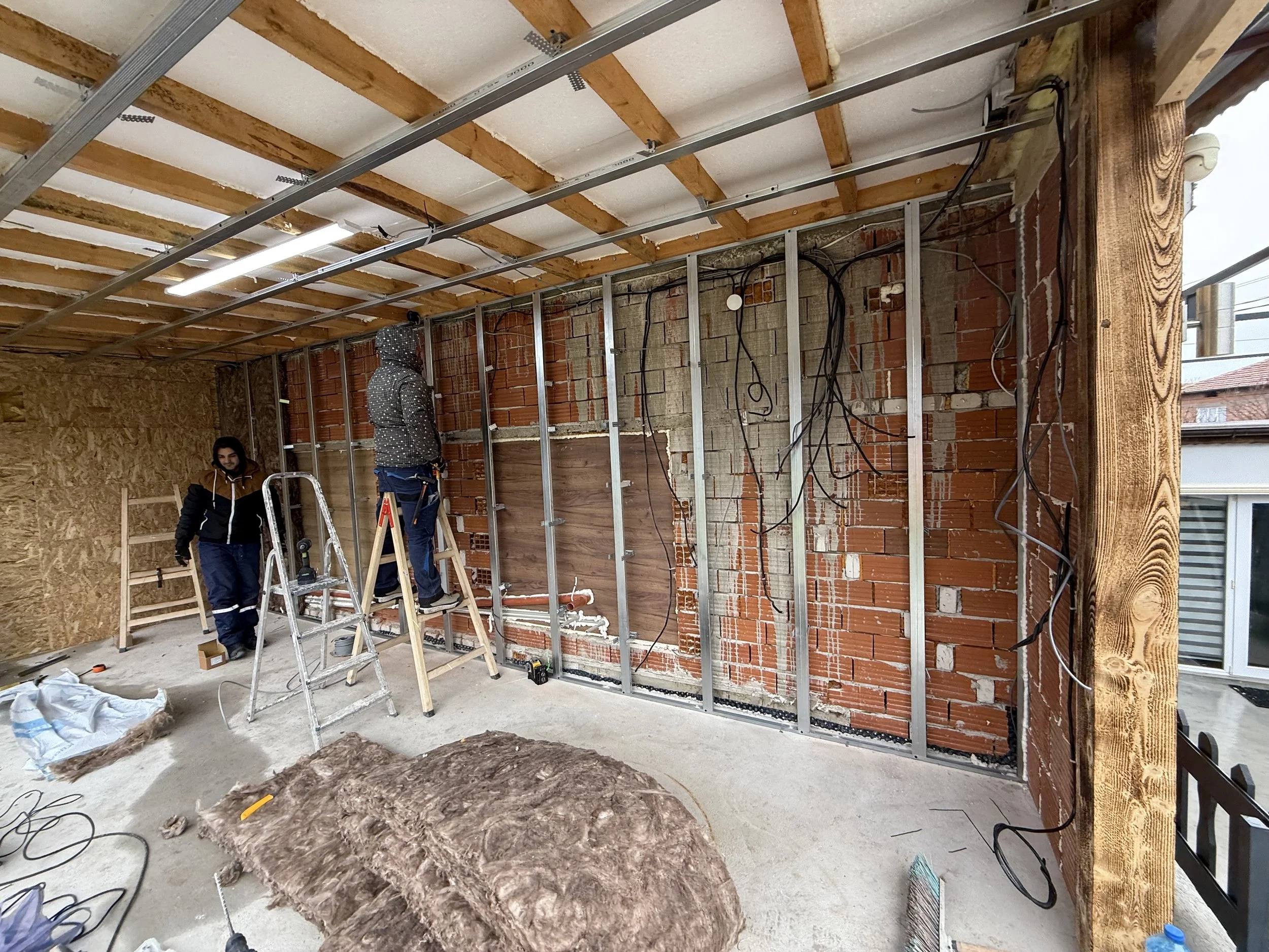 „Двама работници монтират или ремонтират стена в интериорно помещение. Един работник е на стълба, другият държи малка стълба. Стената има видими тухли и електрическо окабеляване, а на пода са разпилени строителни материали и инструменти.