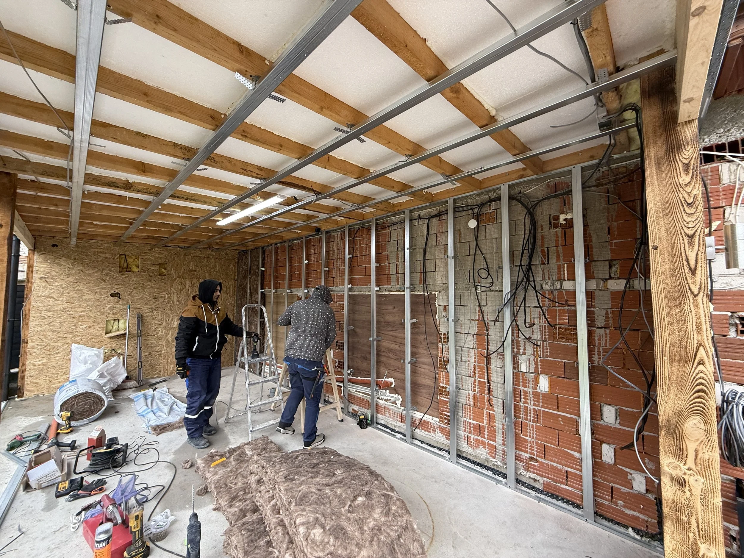 Двама работници монтират метална конструкция на стена в стая в процес на ремонт с видими тухлени стени и електрическо окабеляване. Строителство и довършителни работи с ПДД Констръкшънс в Пловдив