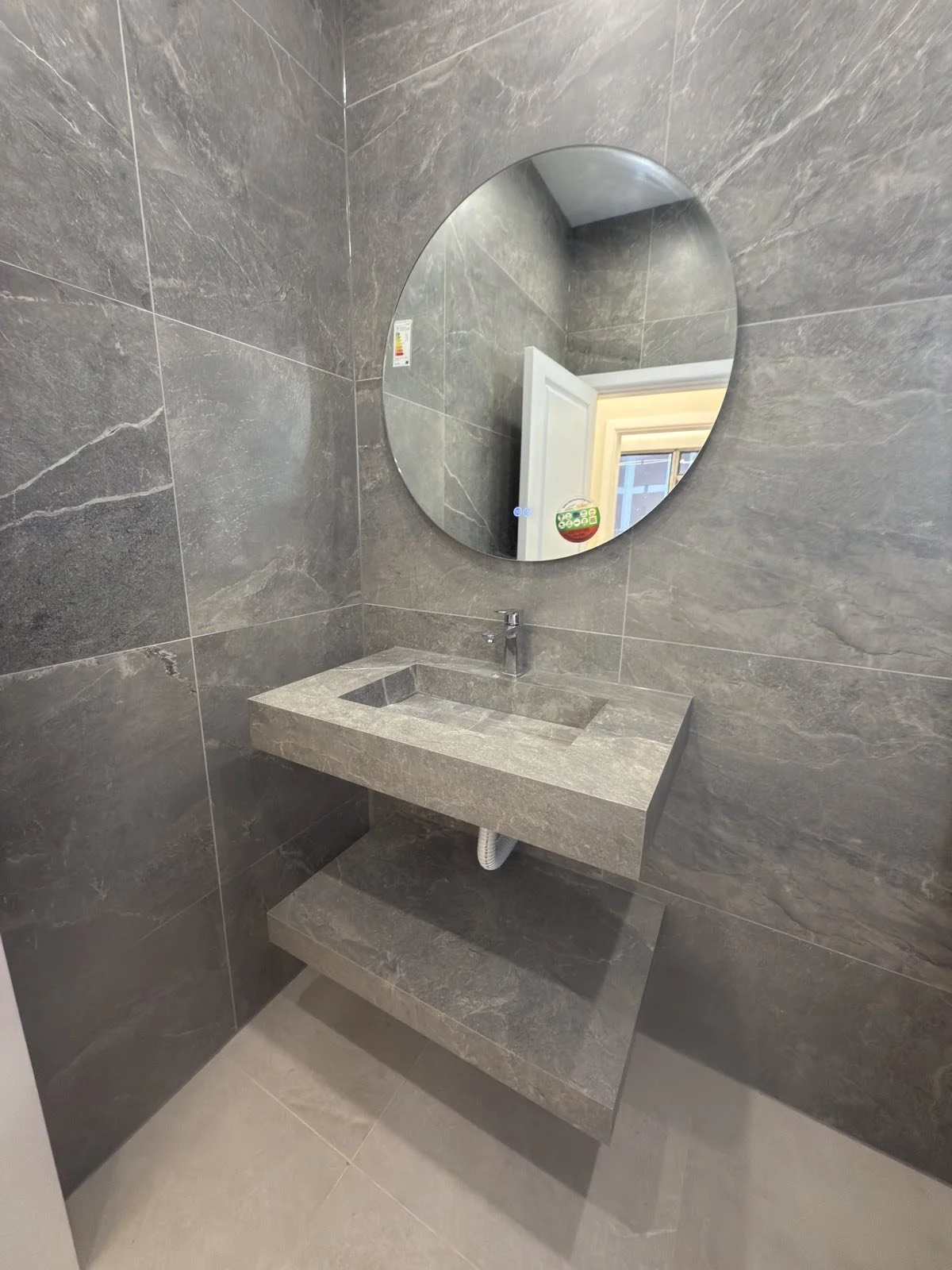 Дизайнерска мивка от лят камък с поръчков плот и кръгло огледало върху широкоформатен сив гранитогрес.