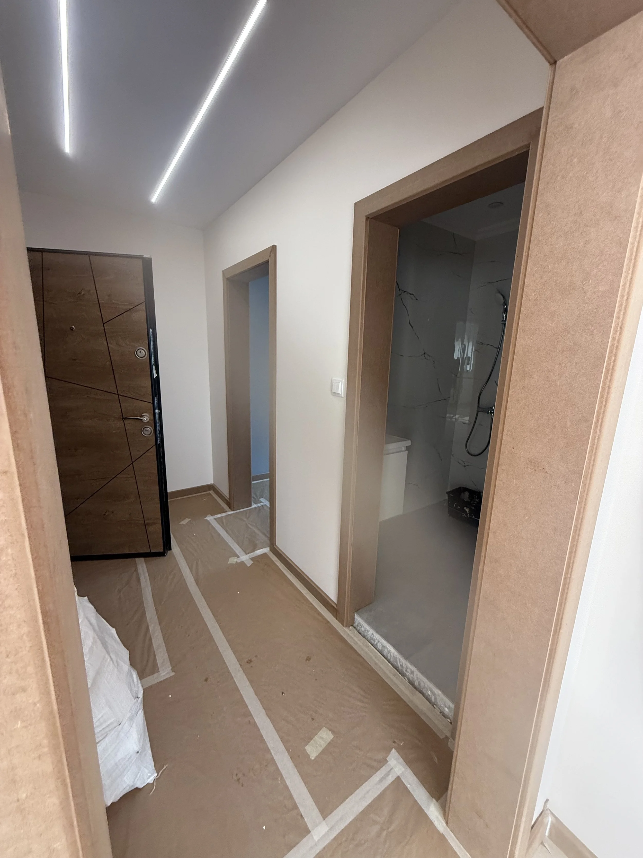 „Вътрешен коридор в Пловдив с две врати – една към стая с мраморни стени и душ, другата към стая със синя стена. Подът и касите са покрити с кафява защитна хартия, а вляво се вижда тъмен дървен шкаф. Таванът има модерно линейно LED осветление – ремон