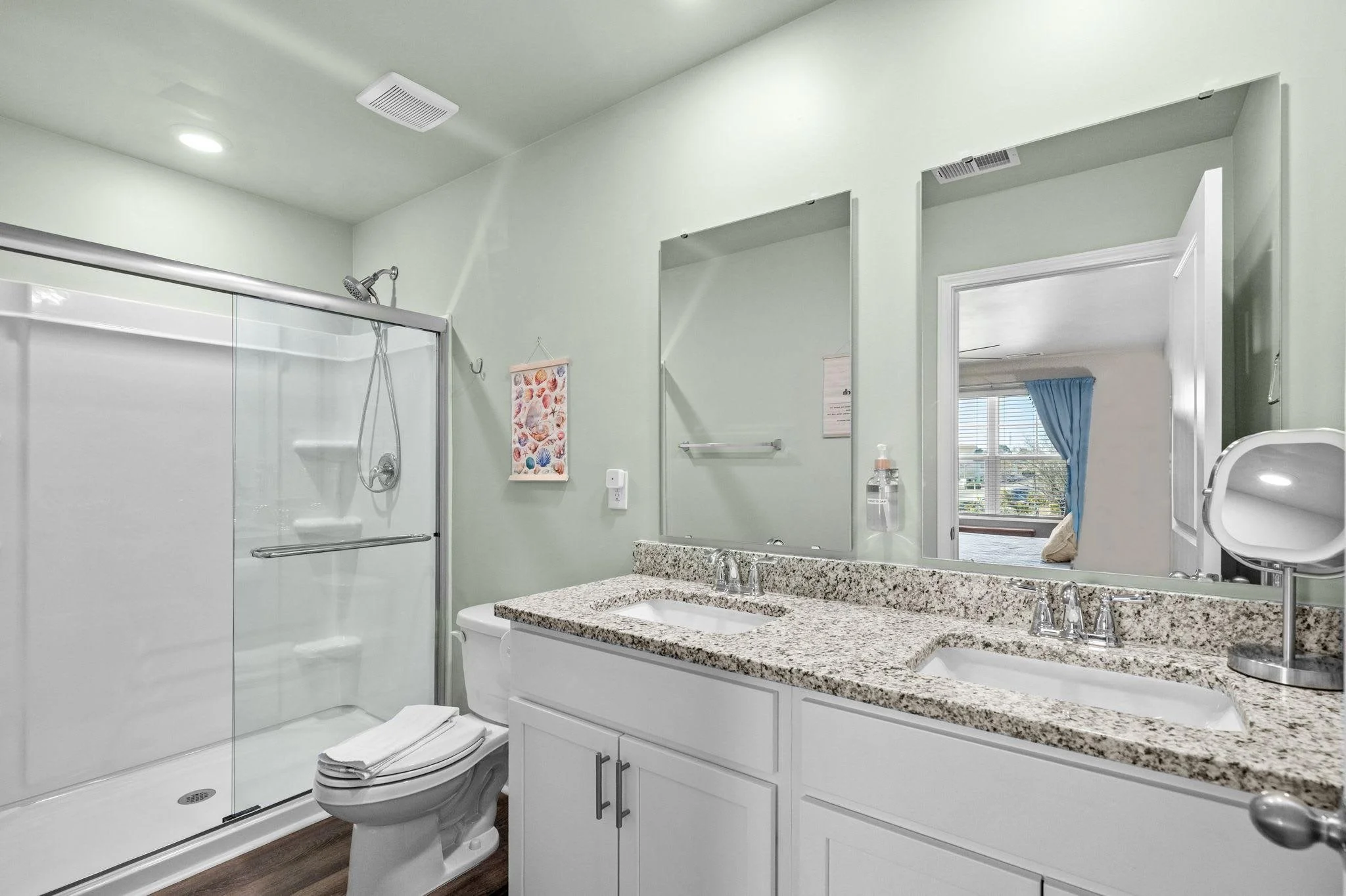 Bathroom with a walk-in shower, white toilet, granite countertop double vanity, large mirror, and a view into adjacent room with blue curtains.