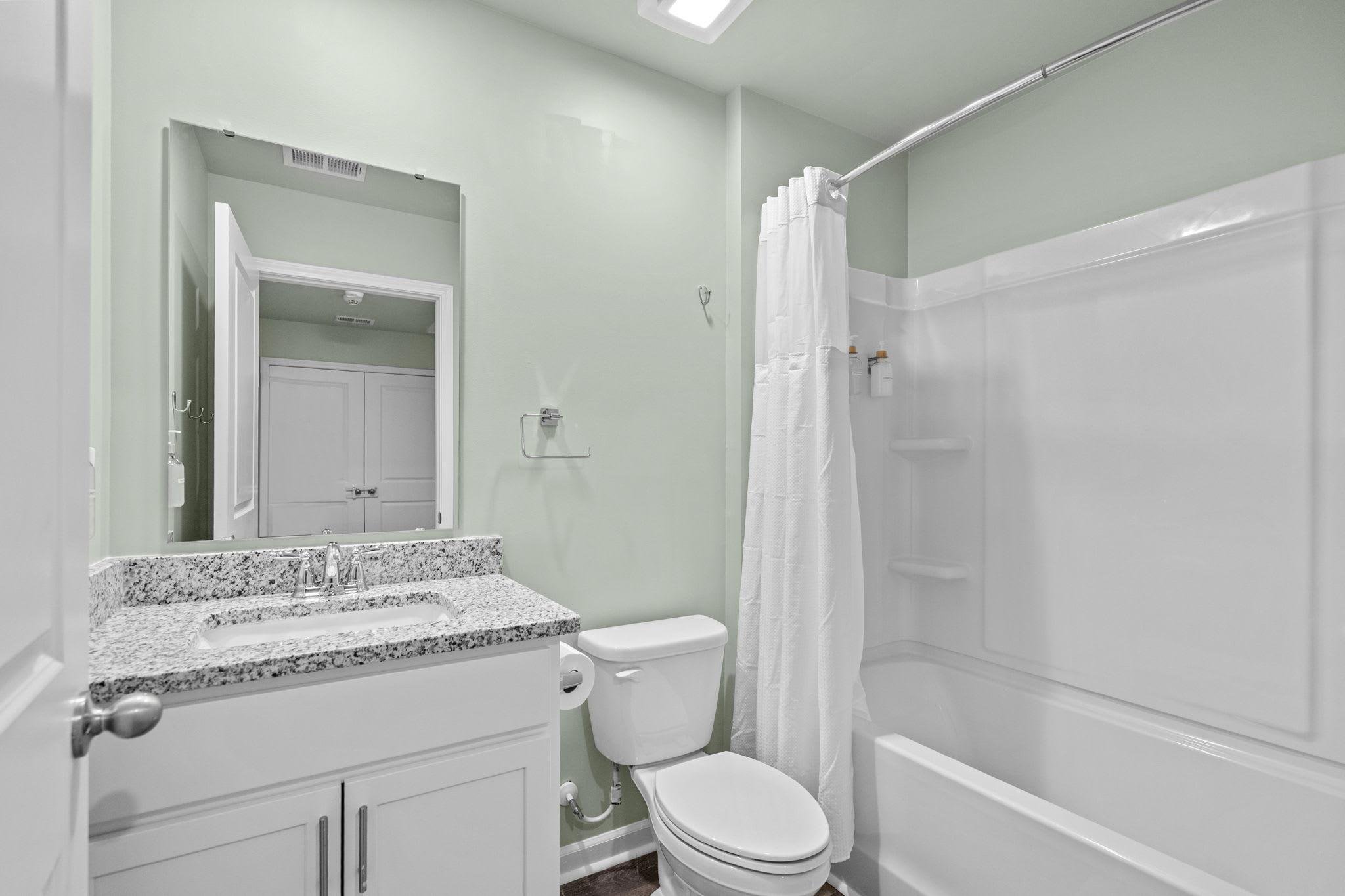 A clean bathroom with light green walls, a white vanity with a granite countertop, a mirror, a toilet, and a bathtub with a shower curtain.