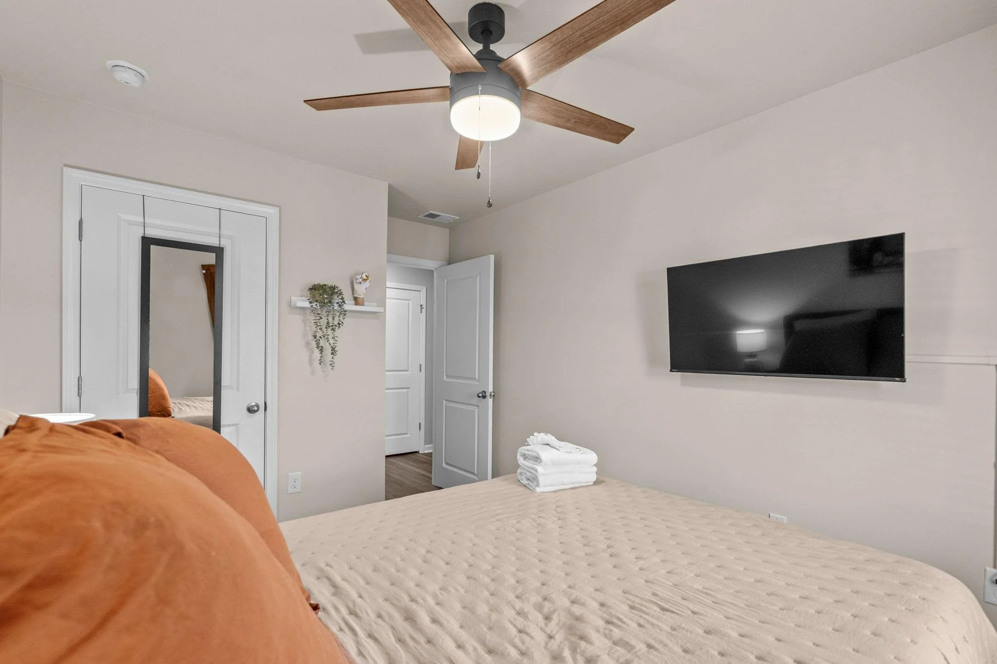 A bedroom with a beige bedspread, stacked white towels, a wall-mounted flat-screen TV, a ceiling fan with wooden blades, and a door leading to another room.