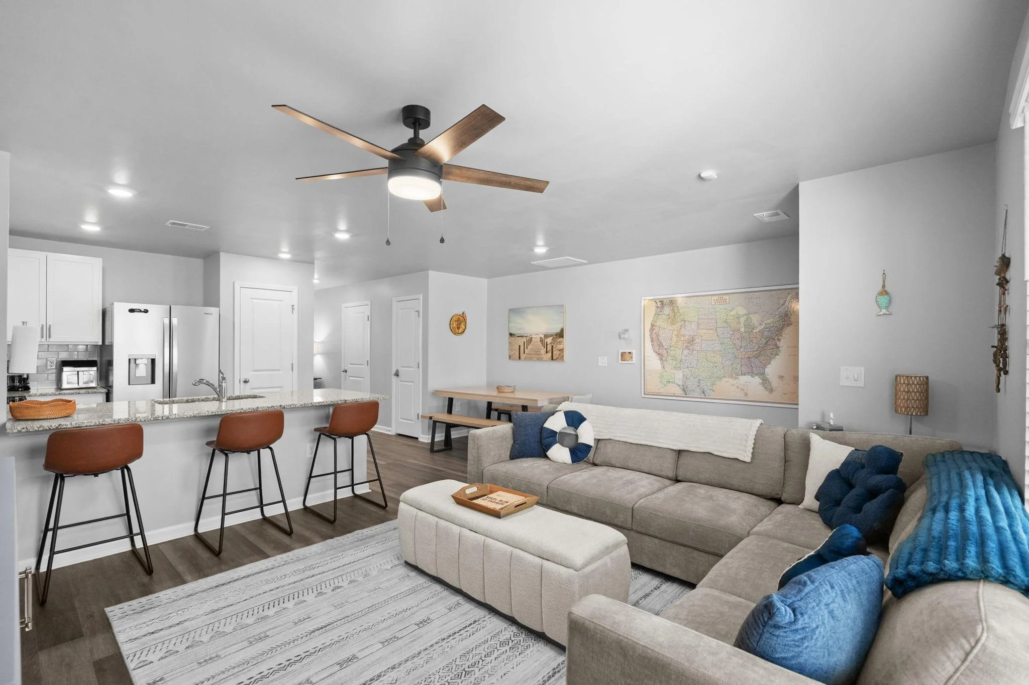 Open-concept living room and kitchen with a ceiling fan, sectional sofa with blue cushions, map on the wall, and kitchen island with three brown bar stools.