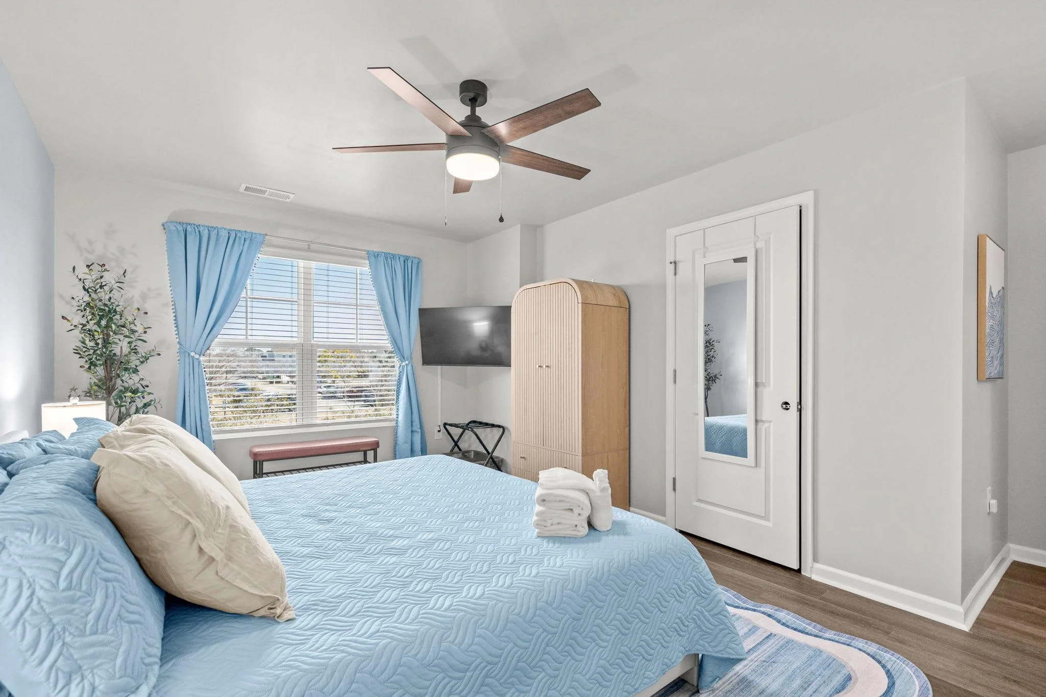 A cozy bedroom with a large bed covered in a blue quilt and beige pillows, a window with blue curtains, a ceiling fan, a matching blue and white rug, a flat-screen TV, a wooden armoire, and a mirror.