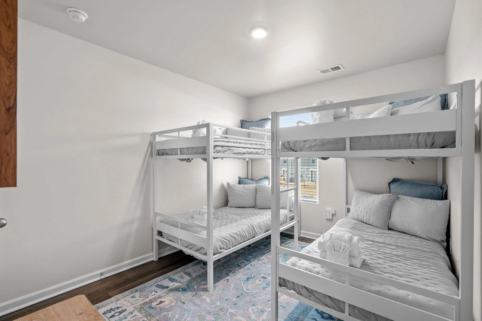 Room with four beds with white frames, two upper and two lower, arranged in a cluster, with pillows and linen, a window in the background, and a colorful area rug on a dark wooden floor.