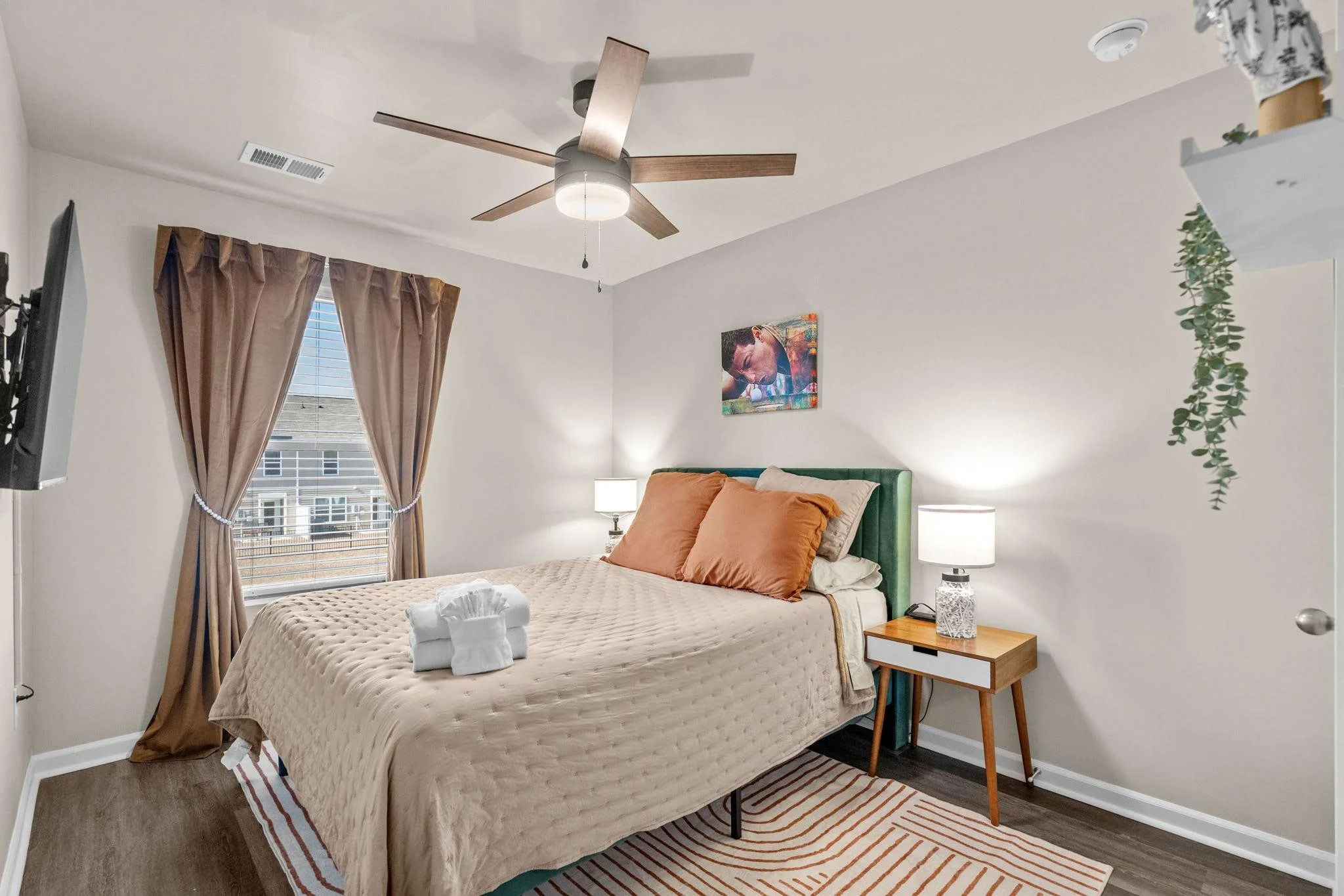 A bedroom with a bed, brown curtains, a window, a ceiling fan, two bedside lamps, a framed picture, and a small wooden nightstand.