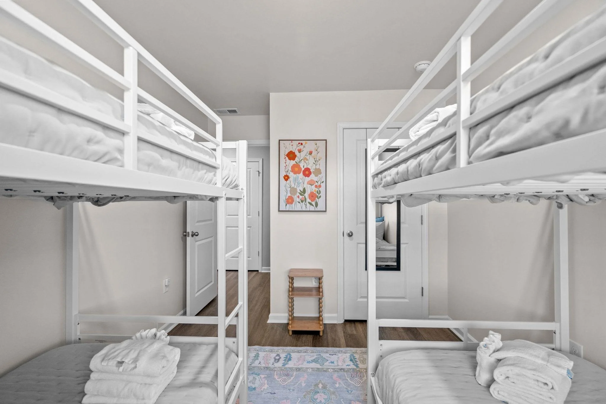 A bedroom with two sets of white bunk beds, with folded towels on each bed, a small wooden step stool, a colorful floral wall art, a mirror on the door, and a rug on a wooden floor.
