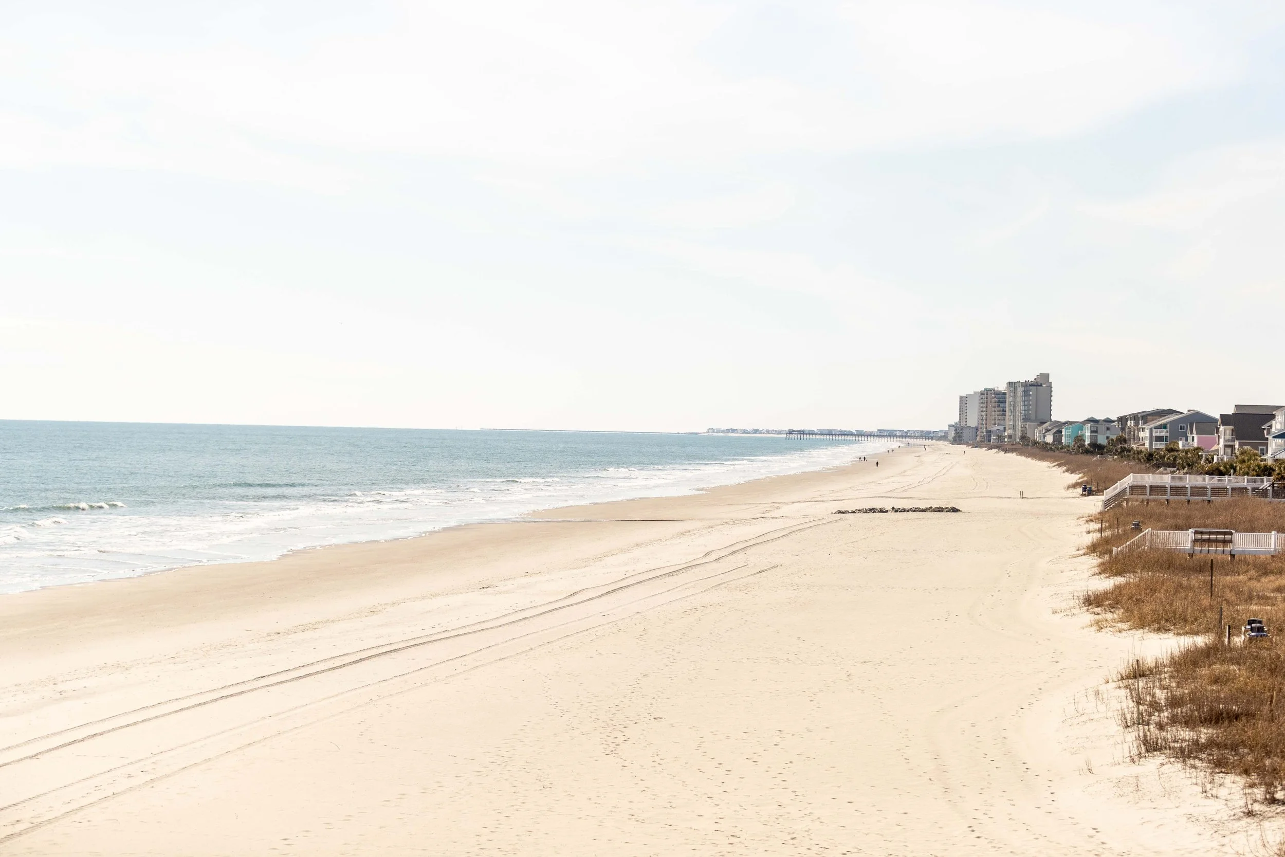 Empty sandy beach with light-colored sand, ocean waves gently lapping the shore, and a line of beachfront houses and condos in the distance under a partly cloudy sky.