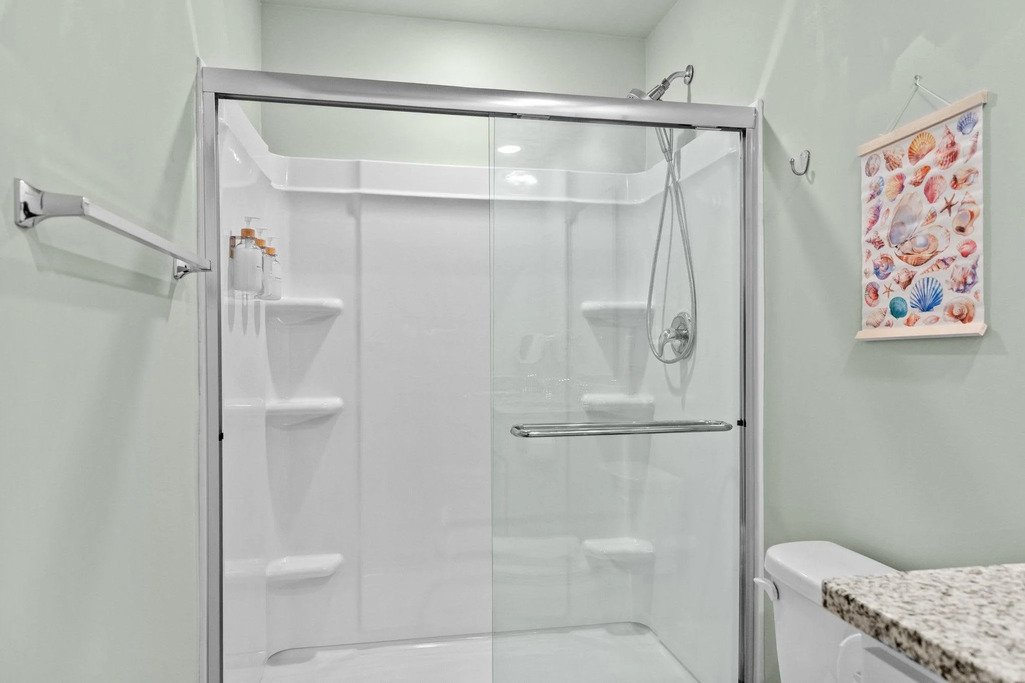 Bathroom with a glass shower enclosure, soap bottles, decorative ocean-themed wall hanging, and a granite countertop.