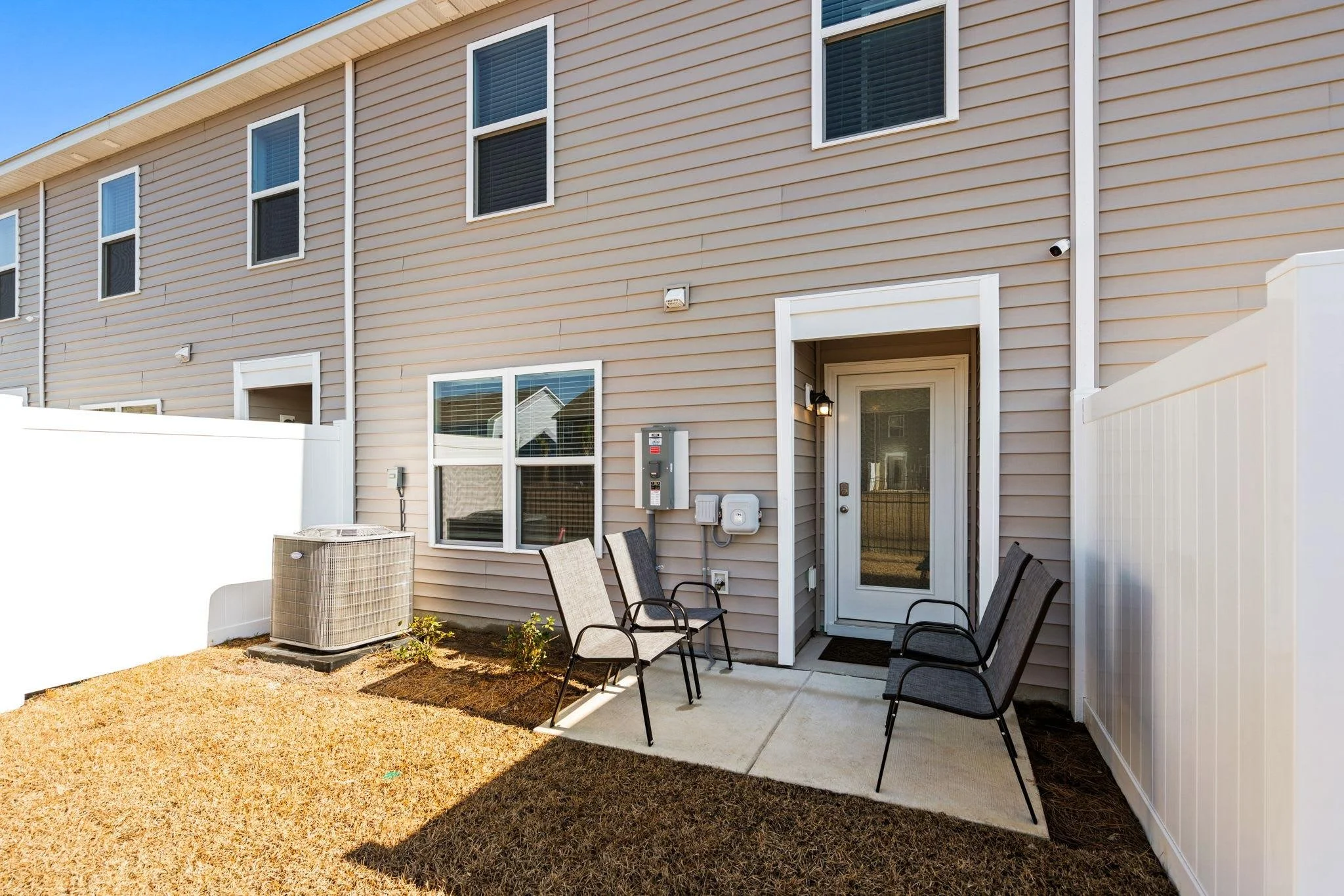 Back patio area with two chairs, a small patch of grass, and an air conditioning unit outside a beige townhouse with a white privacy fence.