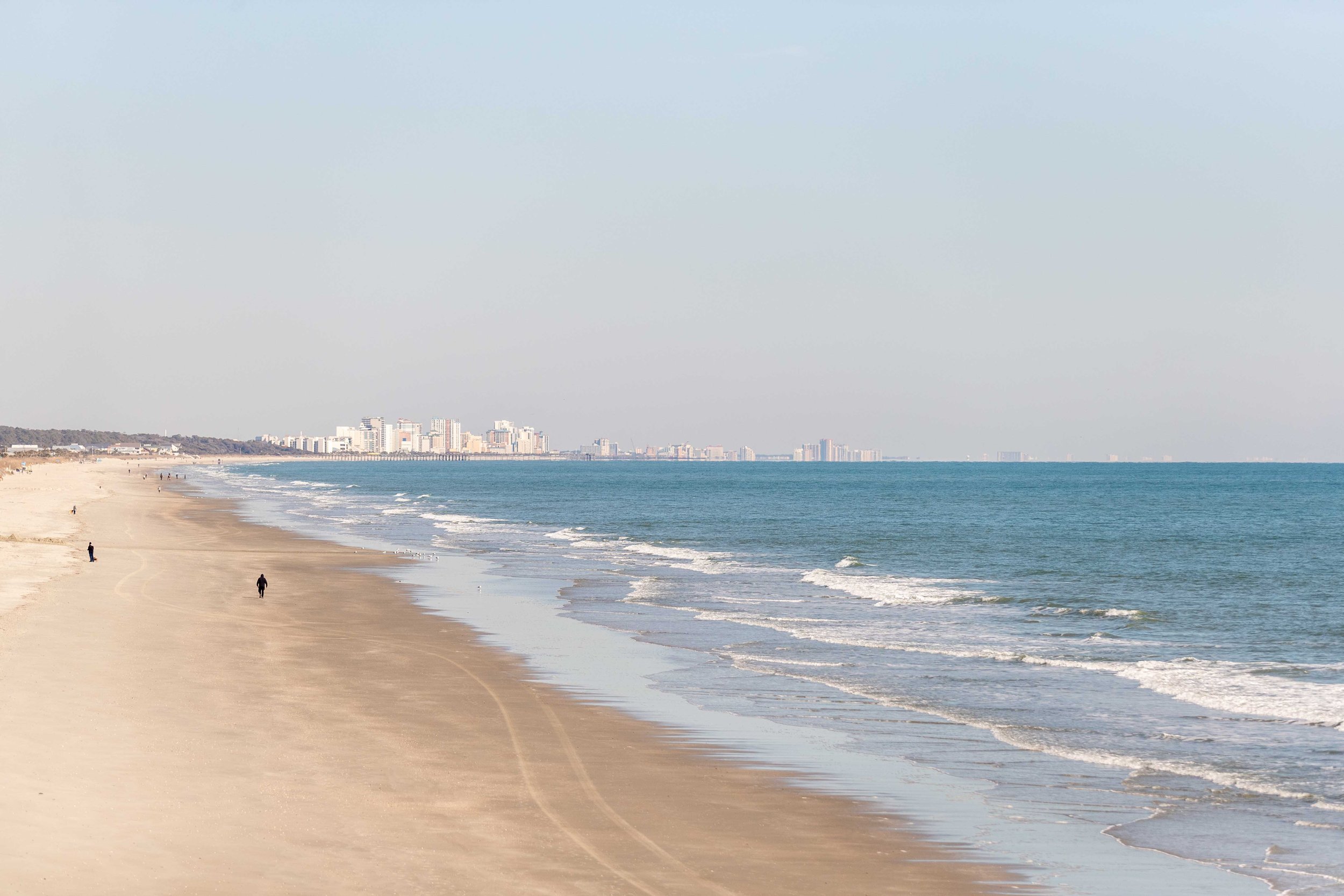 A stretch of sandy beach with a few people walking, an ocean with gentle waves, and a distant skyline of high-rise buildings under a clear sky.