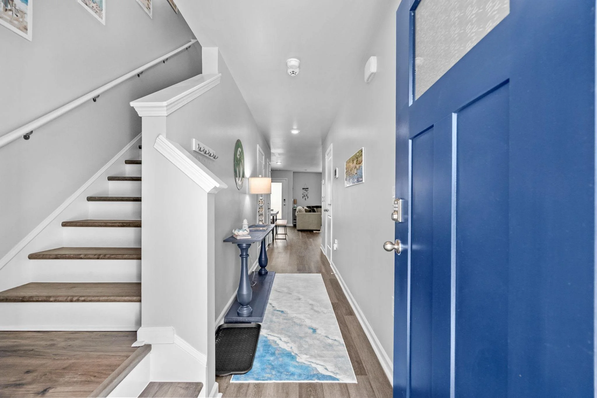 View of a home's entryway with a blue front door, white walls, wooden staircase, a hallway with colorful wall art, and a living room in the background.