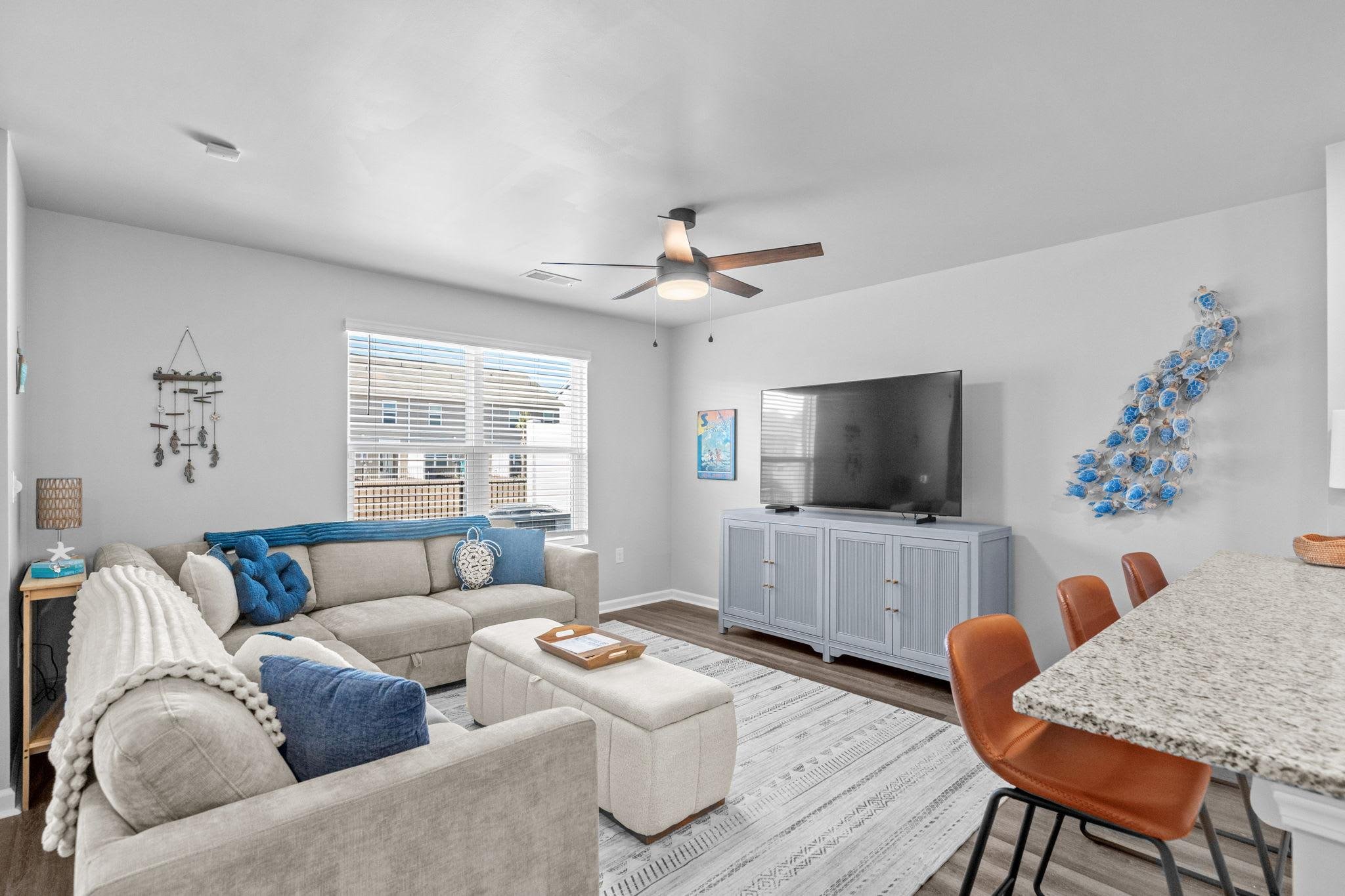 Living room with beige sofa, blue pillows, white and beige ottoman, gray TV stand with a large flat-screen TV, window with blinds, wall art, ceiling fan, patterned rug, and bar stools at a granite kitchen counter.