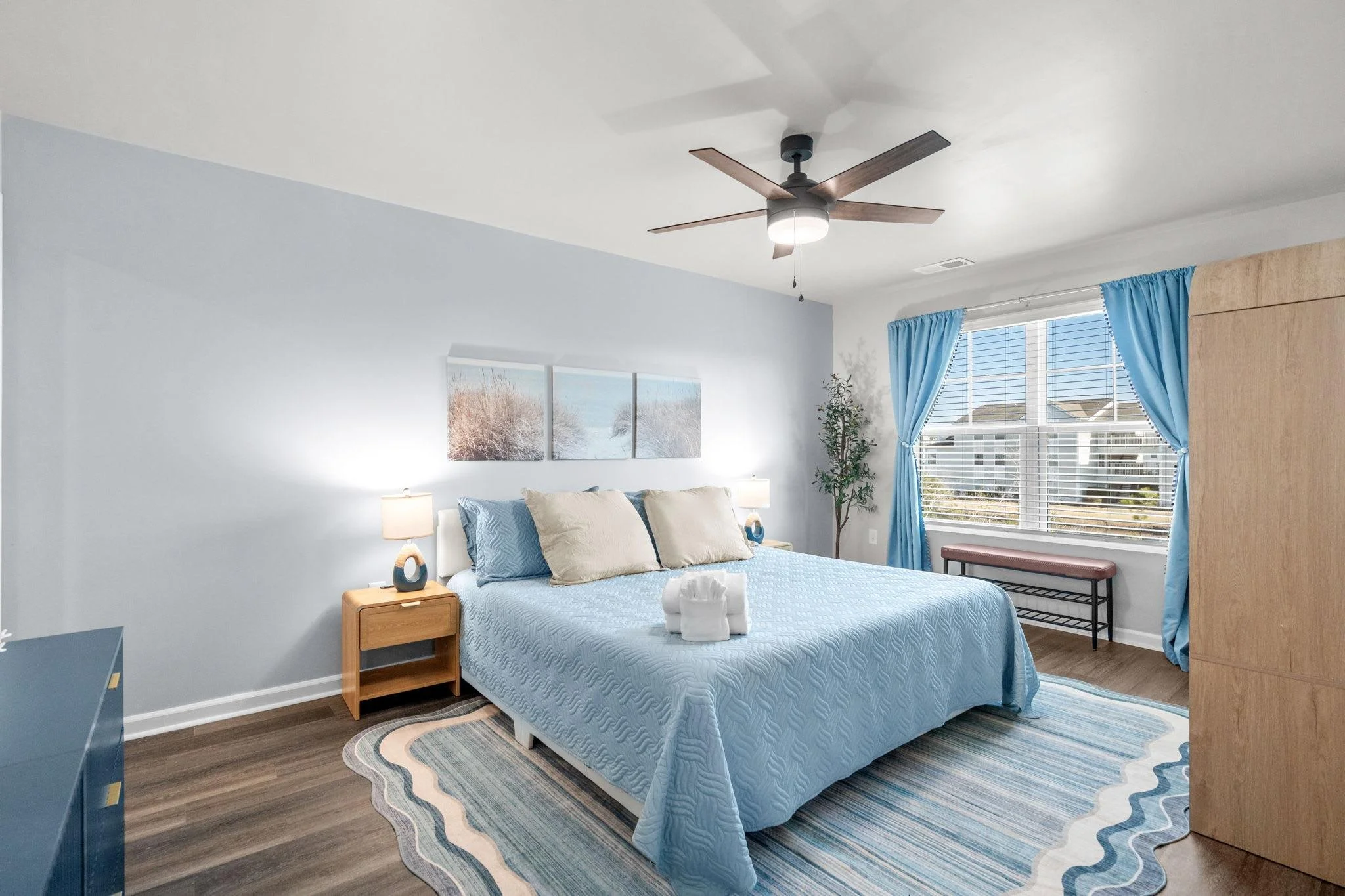 A bright bedroom with a large bed covered in a light blue quilt and beige pillows, flanked by wooden nightstands with lamps. There is a large window with blue curtains, a bench under the window, a potted plant in the corner, and a ceiling fan with wo
