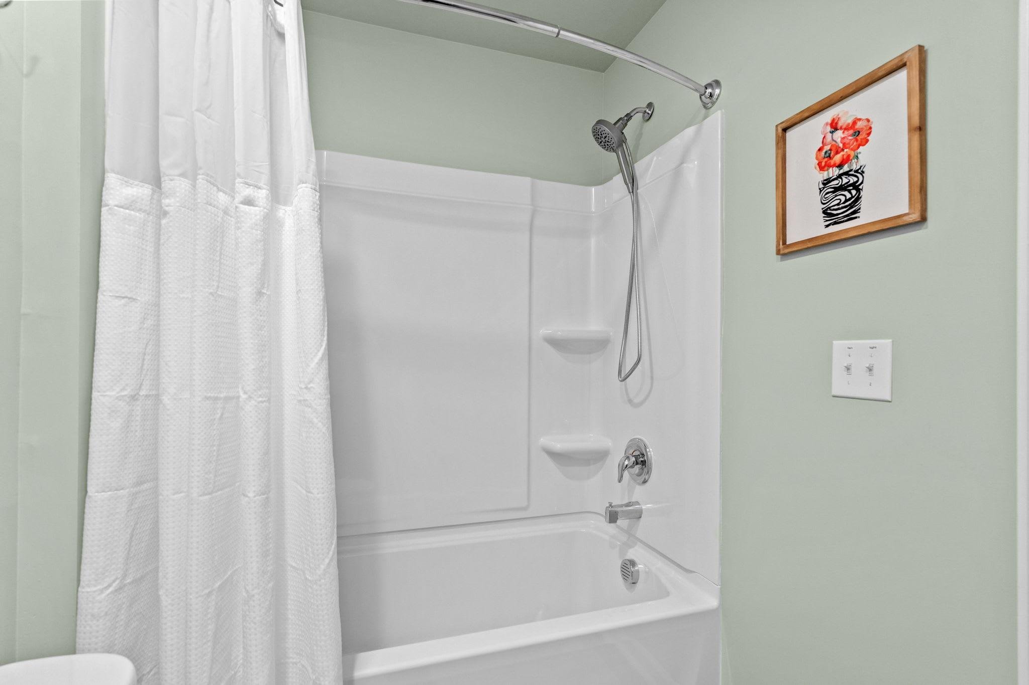 A bathroom with a white bathtub and shower, white shower curtain, green walls, a framed floral artwork, and a light switch.