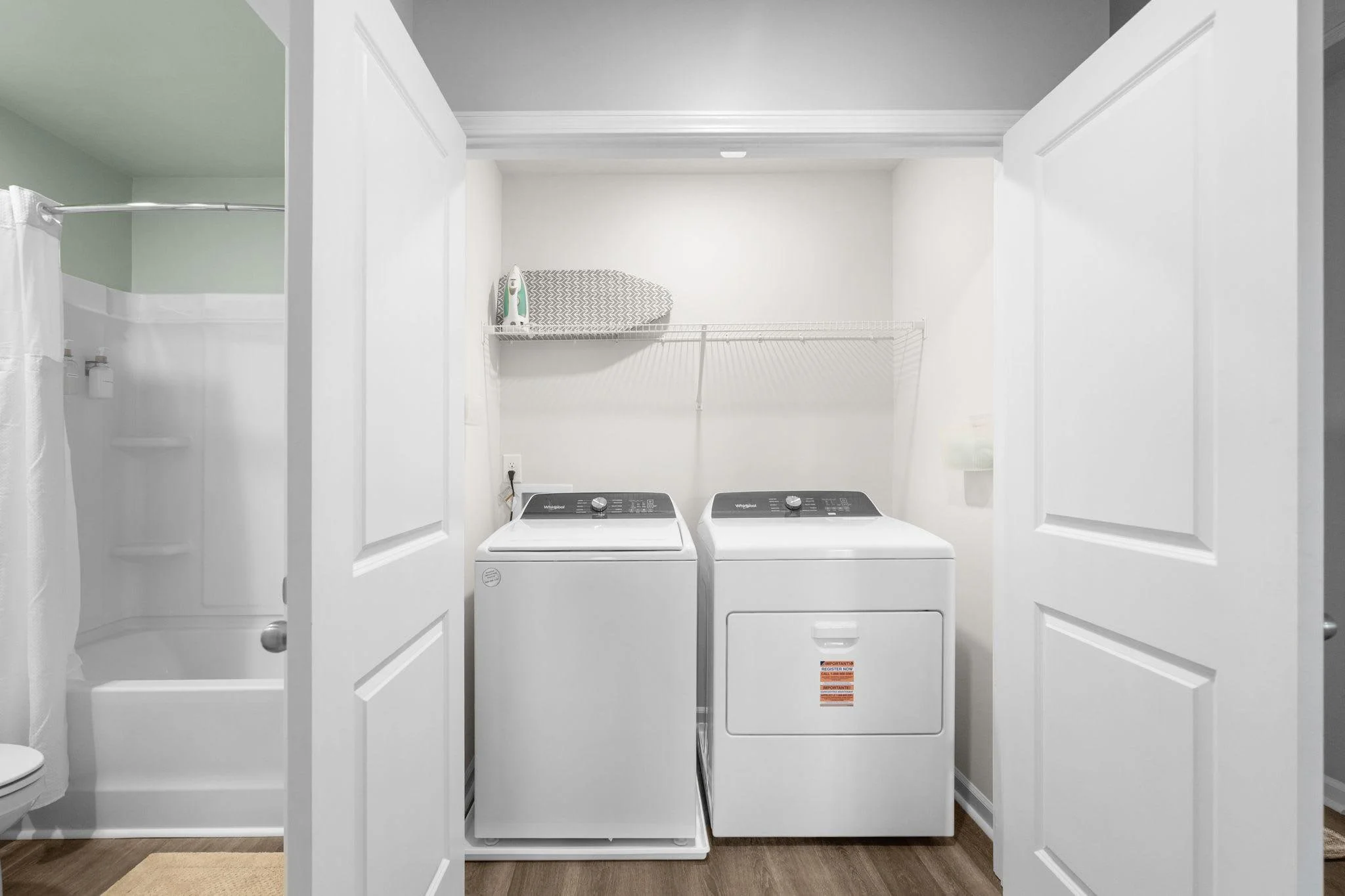 Laundry room with a washing machine and dryer, white walls, wire shelf with iron and iron board, and a bathroom with a shower and toilet visible through an open door.