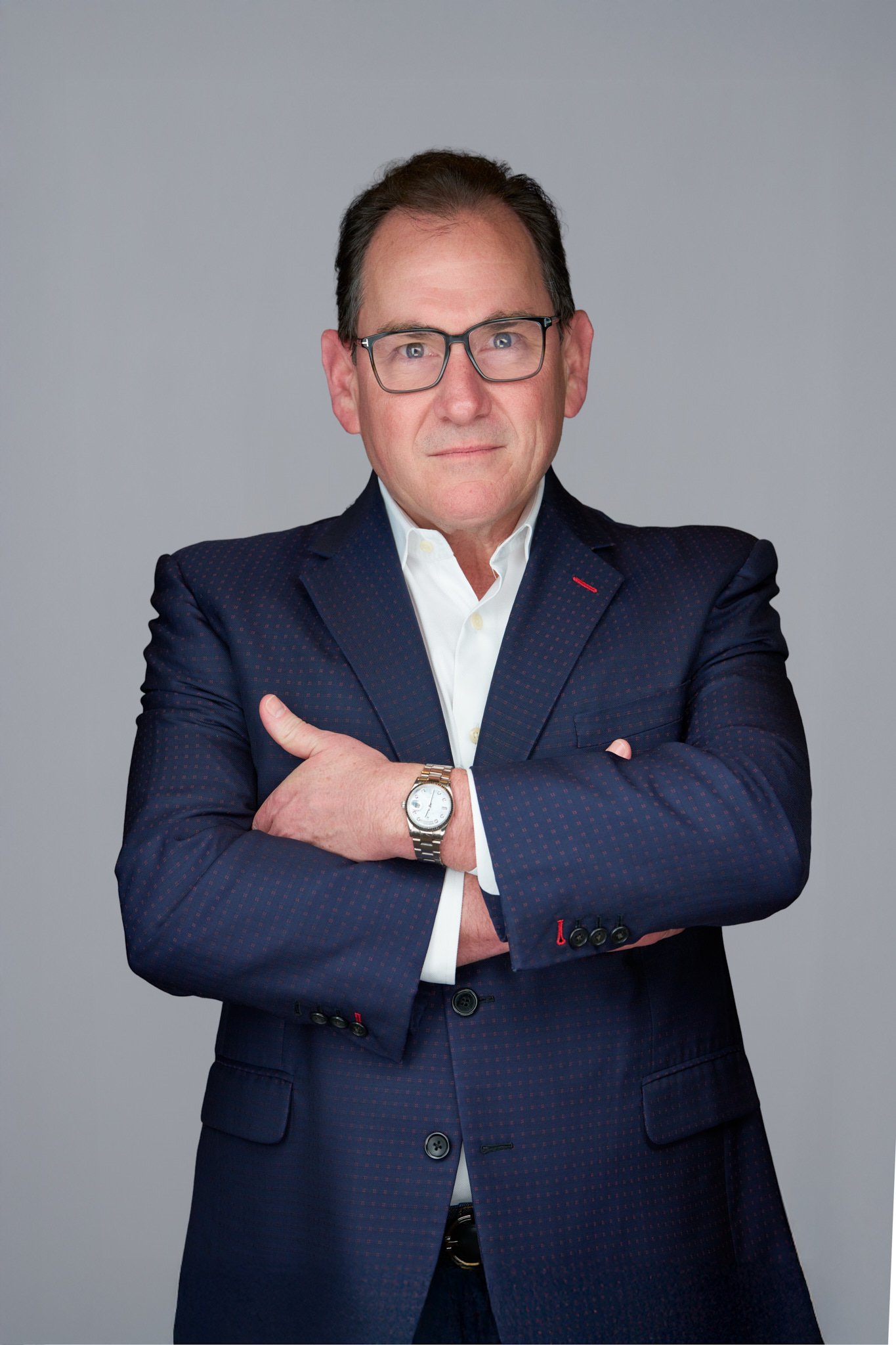 A man with glasses and dark hair, wearing a navy blue suit with a white dress shirt, posing against a plain light gray background with his arms crossed.