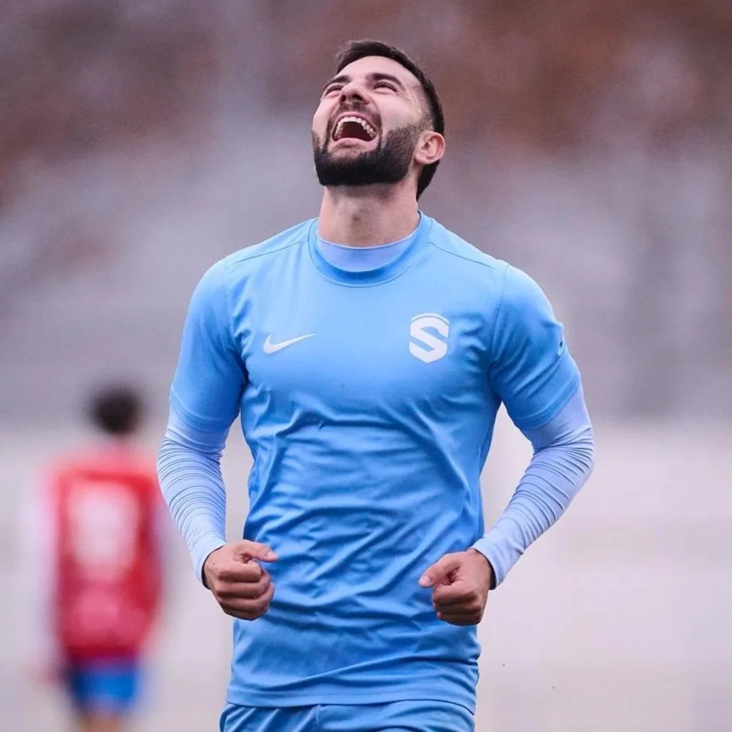 Un hombre con camiseta azul, con barba y cabello corto, celebra con una expresión de felicidad en un campo de fútbol.