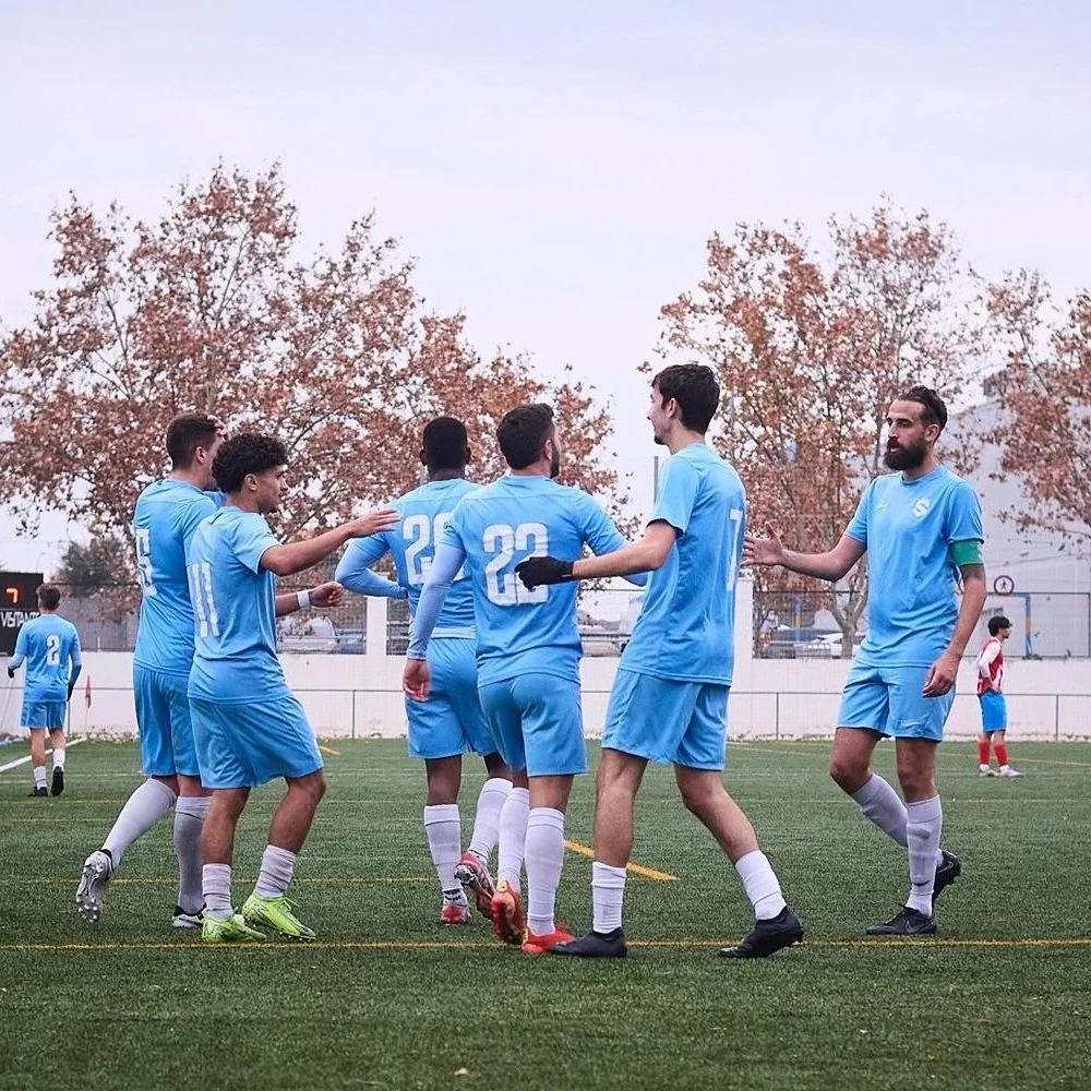 Jugadores de fútbol en un campo, celebrando un gol, vistiendo uniformes azules y en un día nublado.