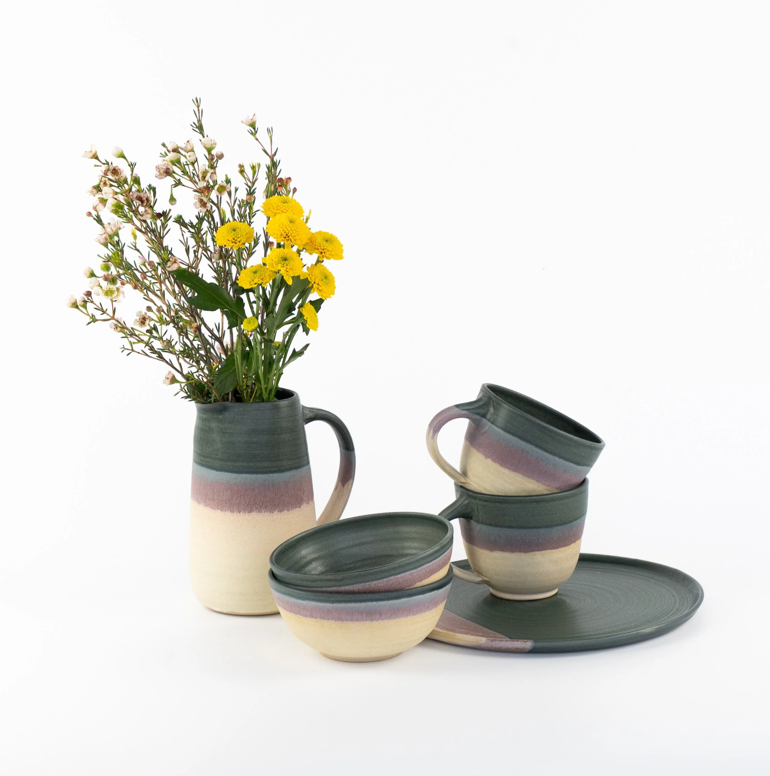 Mehrere handgefertigte Keramikbecher und -krüge in Pastellfarben auf einem Holzregal, mit grünem Blattpflanze im Hintergrund, bei einem Markt im Freien.