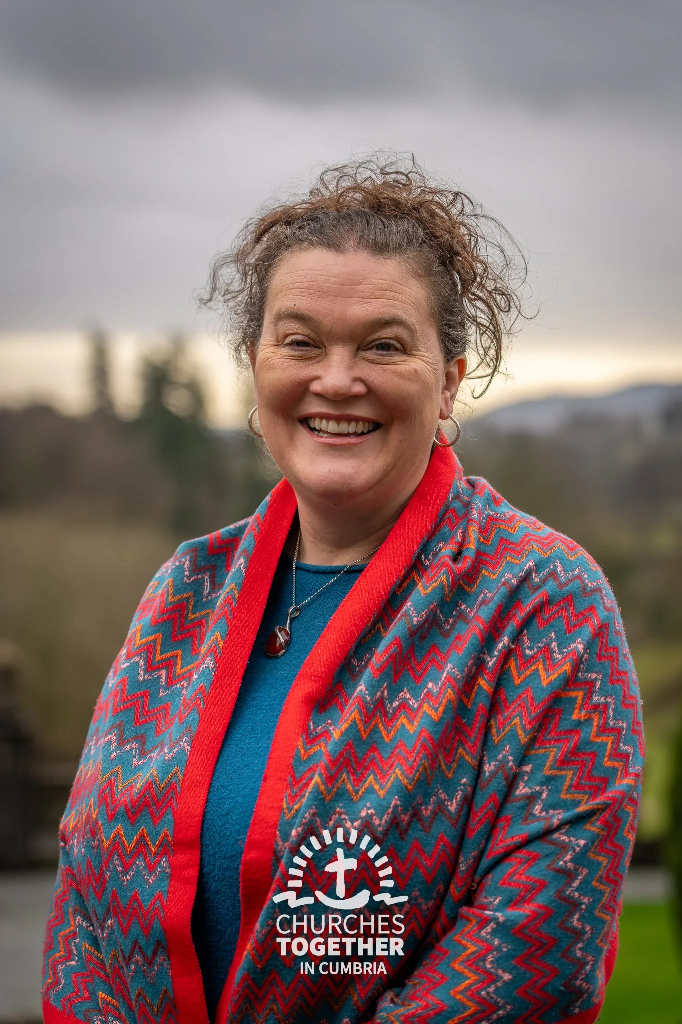 Smiling woman with shoulder-length black hair wearing a blue blouse in a botanical setting with green foliage.