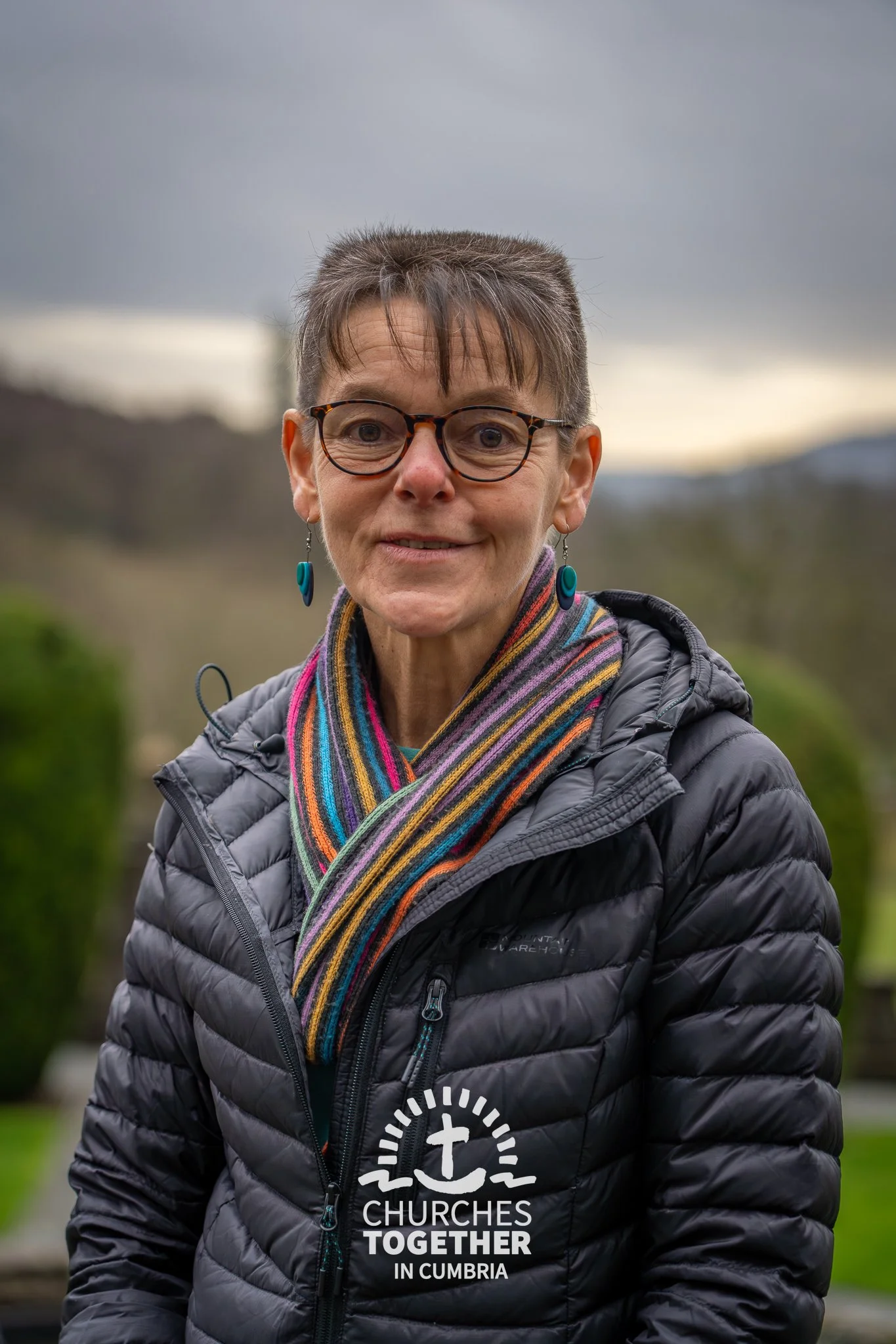 A smiling woman with glasses and curly hair wearing a striped shirt, layered necklaces, and earrings.