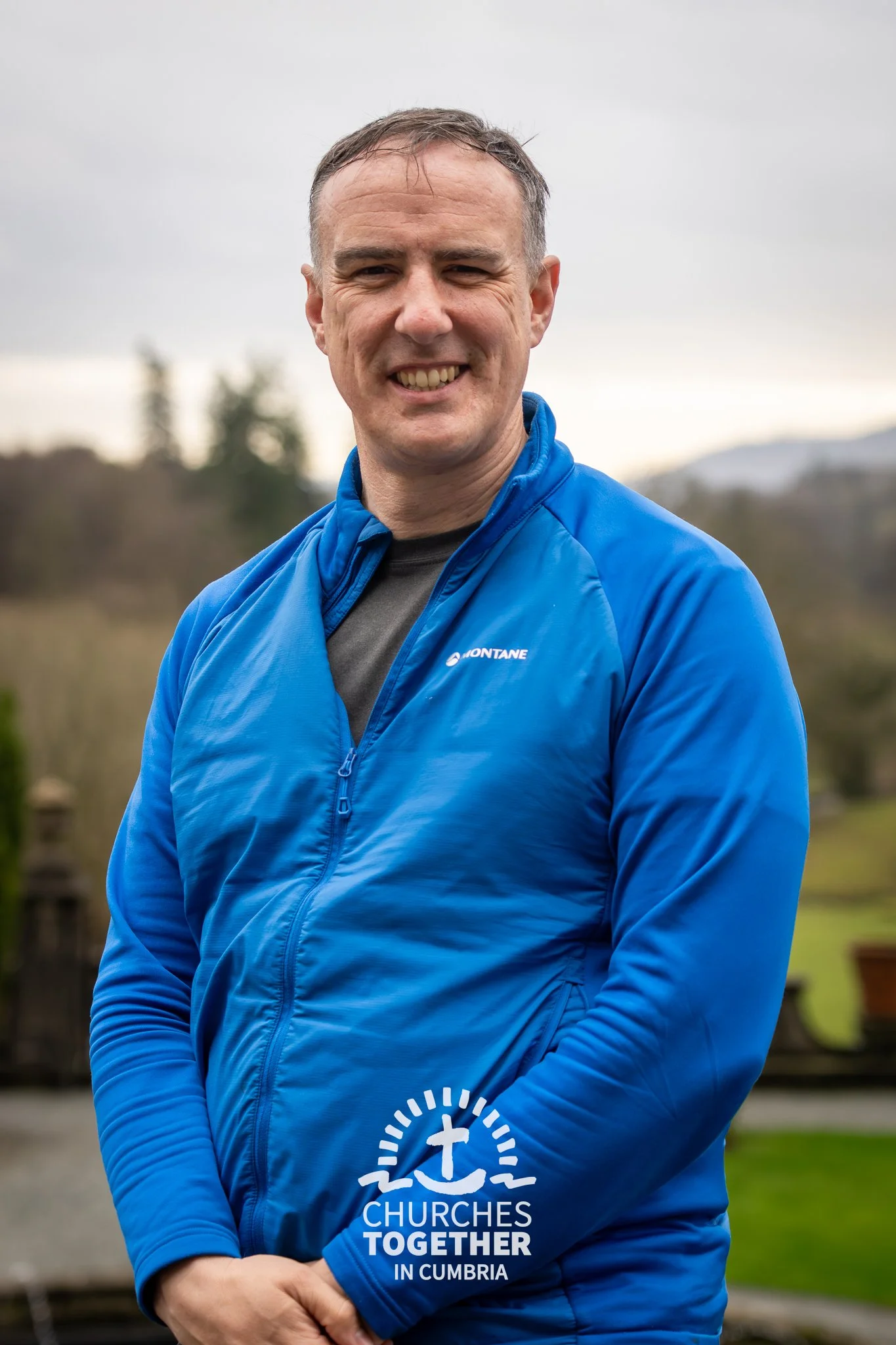 A man with curly dark hair, glasses, and a beard, wearing a dark sweater, sitting indoors with a neutral expression, a plant in the background.