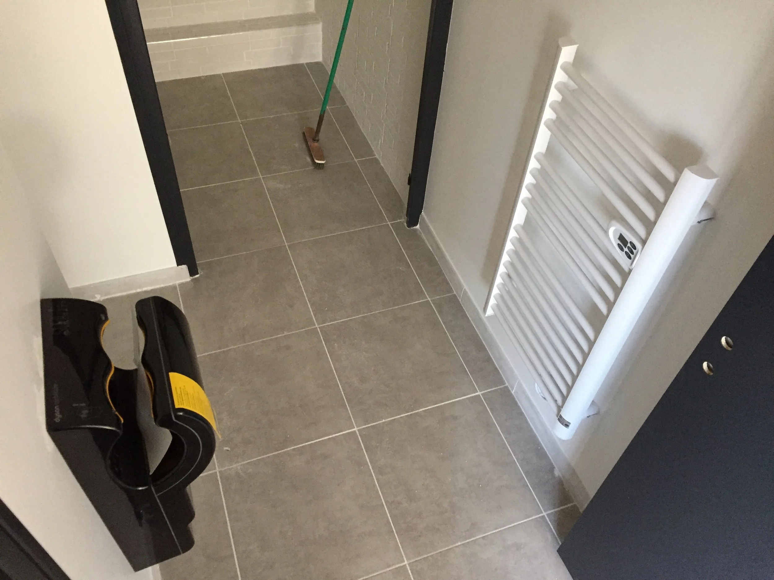 Intérieur d'une pièce avec carrelage gris, un radiateur blanc fixé au mur, un rouleau de papier ou un film protecteur sur le radiateur, un balai en position verticale dans un coin, une station de détection automatique de savon ou de désinfectant mura