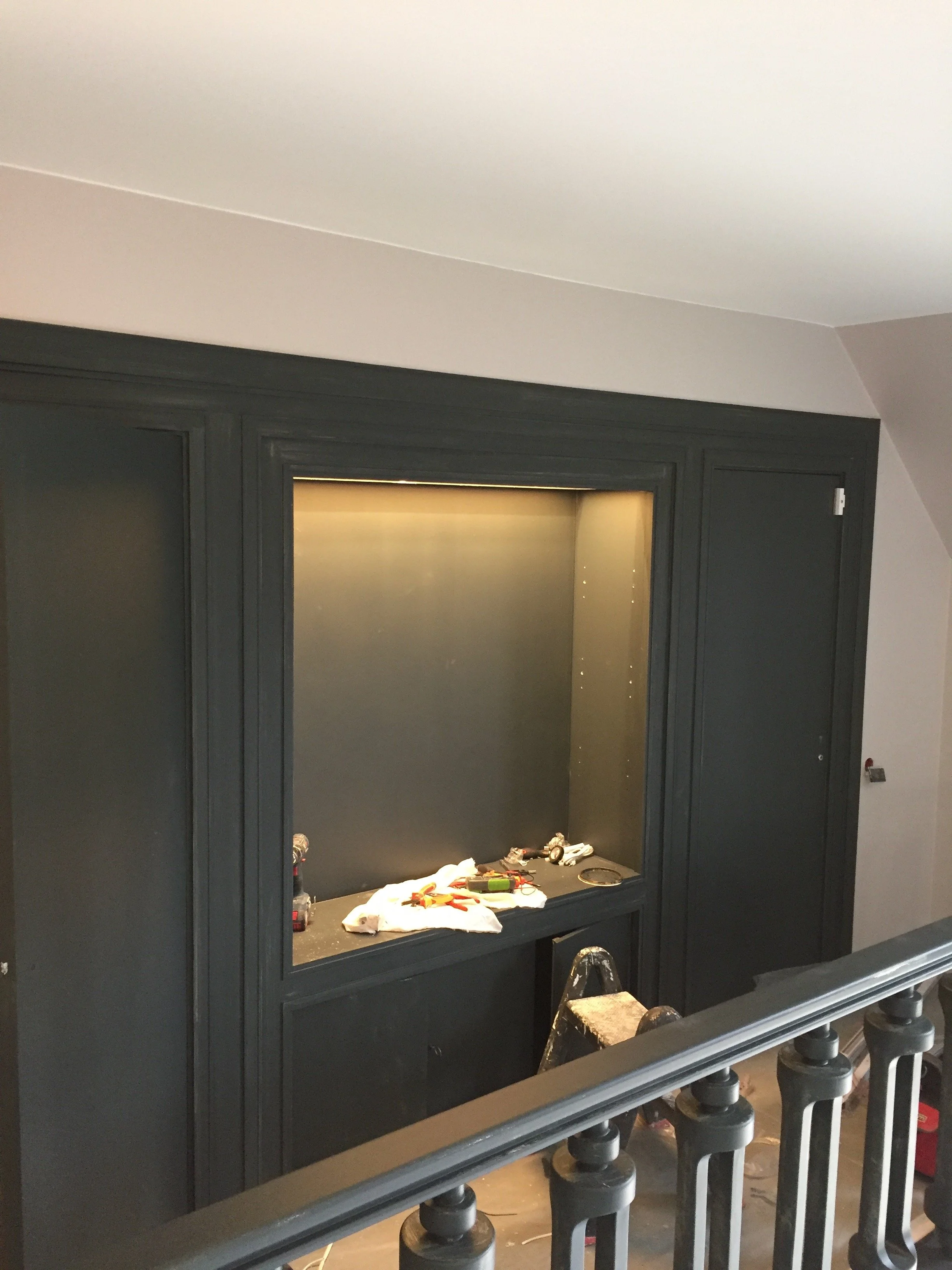 Une pièce en cours de rénovation avec un mur encadré prêt à accueillir une télévision ou un autre écran, des outils et matériaux de bricolage posés sur le rebord, et une escadre en dessous.