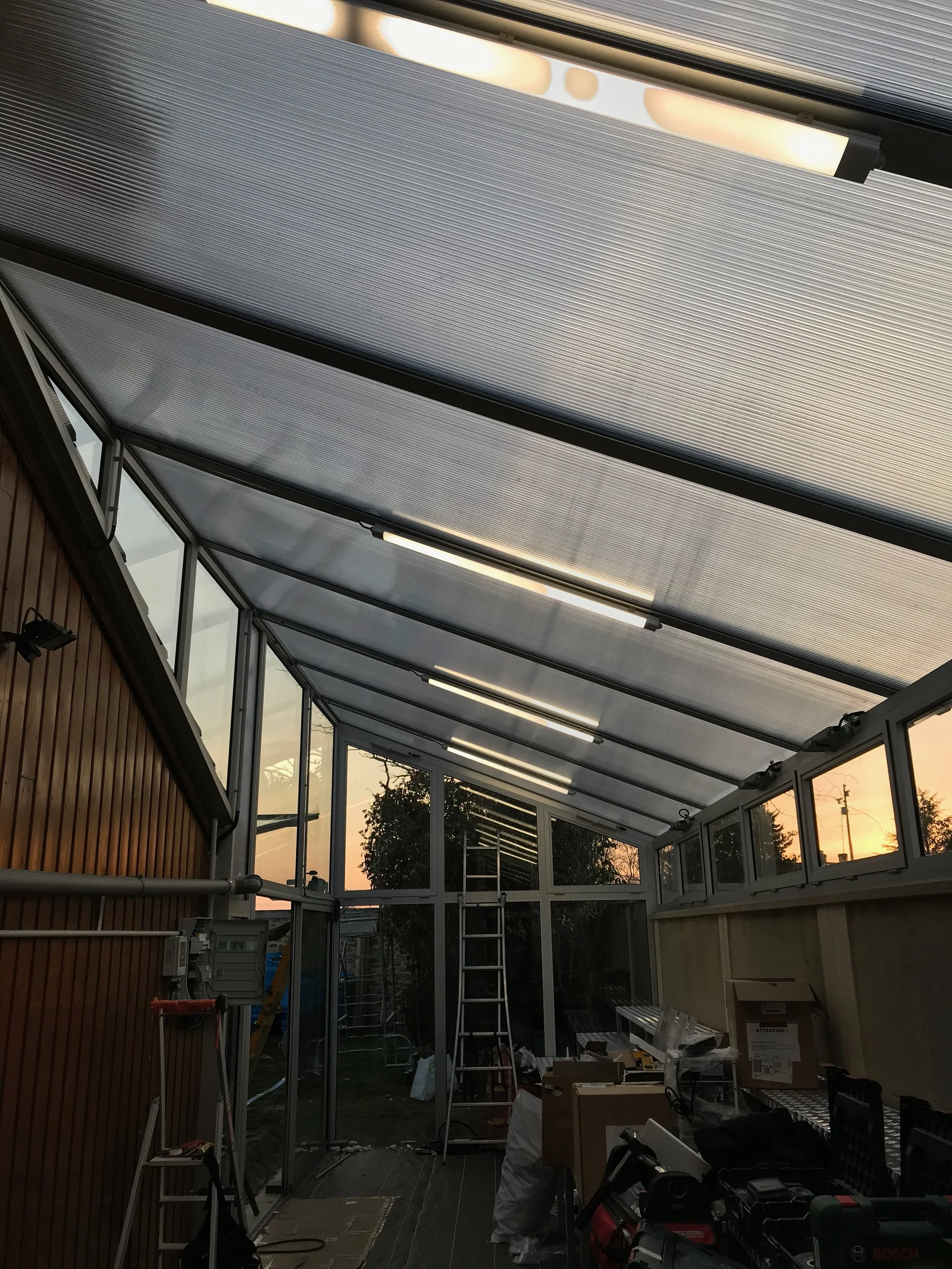 Une véranda en construction avec des outils, escabeau et cartons, le ciel au coucher du soleil visible à travers les fenêtres.