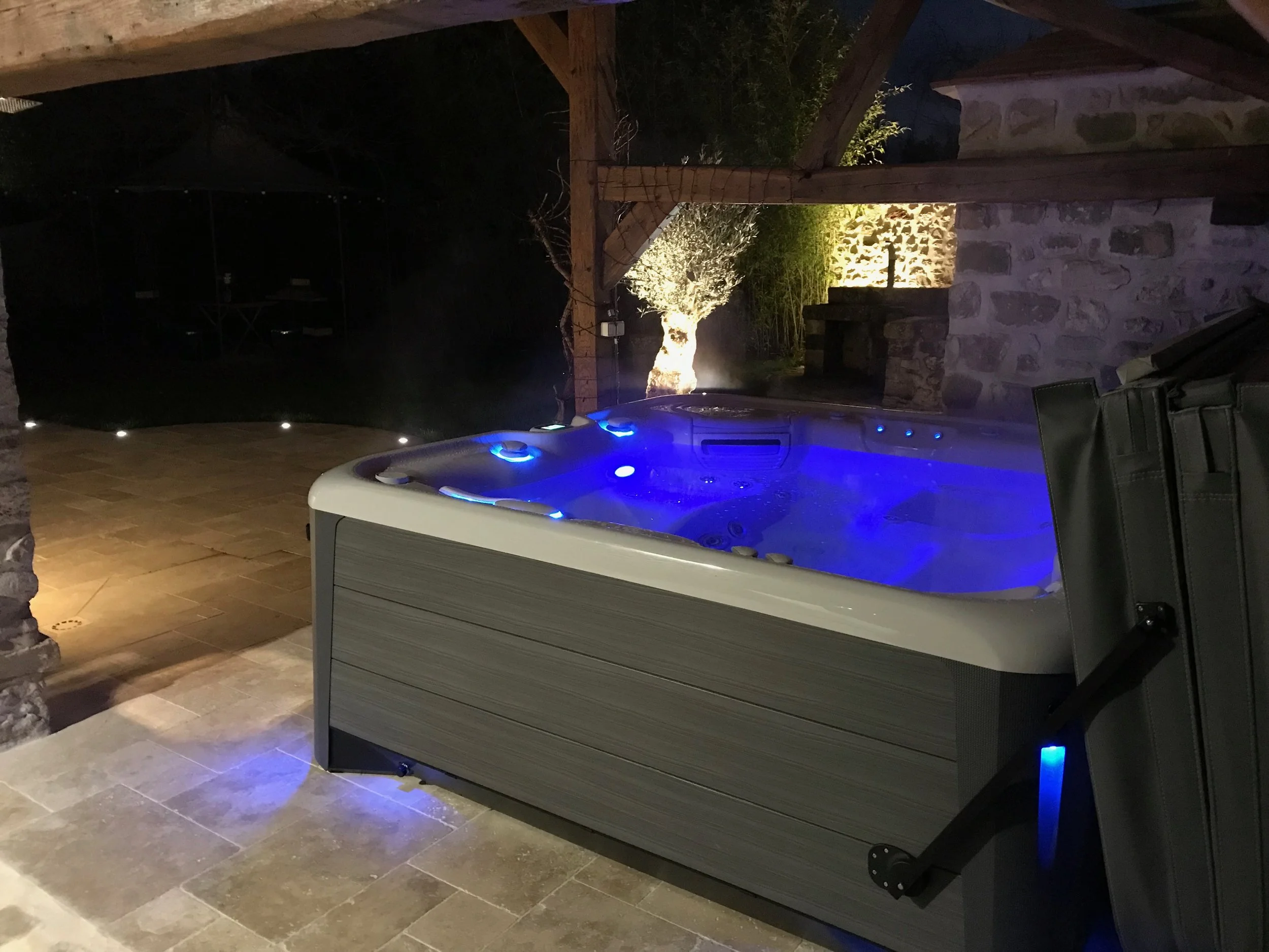 Jacuzzi extérieur éclairé en bleu dans un espace de nuit avec une pergola en bois, des murs en pierre, un arbre éclairé et un sol en carreaux de pierre.