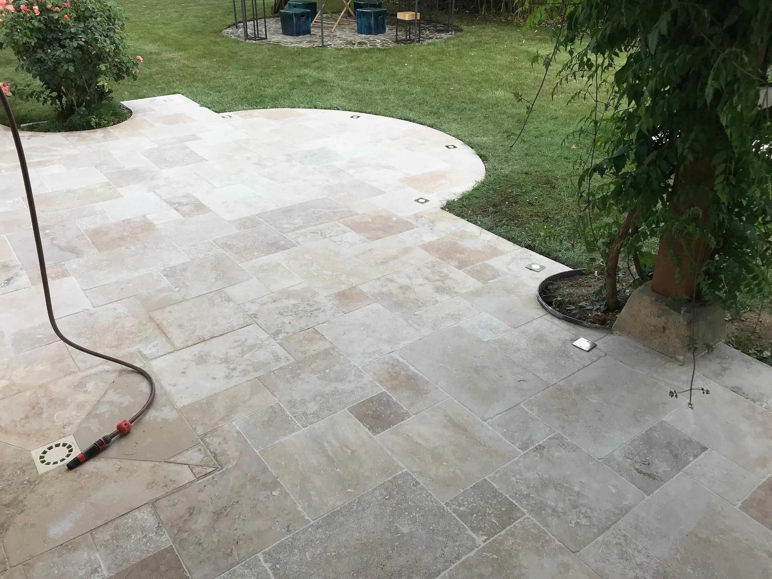 Terrasse en pierre claire avec éclairages encastrés, jardins avec buissons, arbres, et mobilier de jardin en fond.
