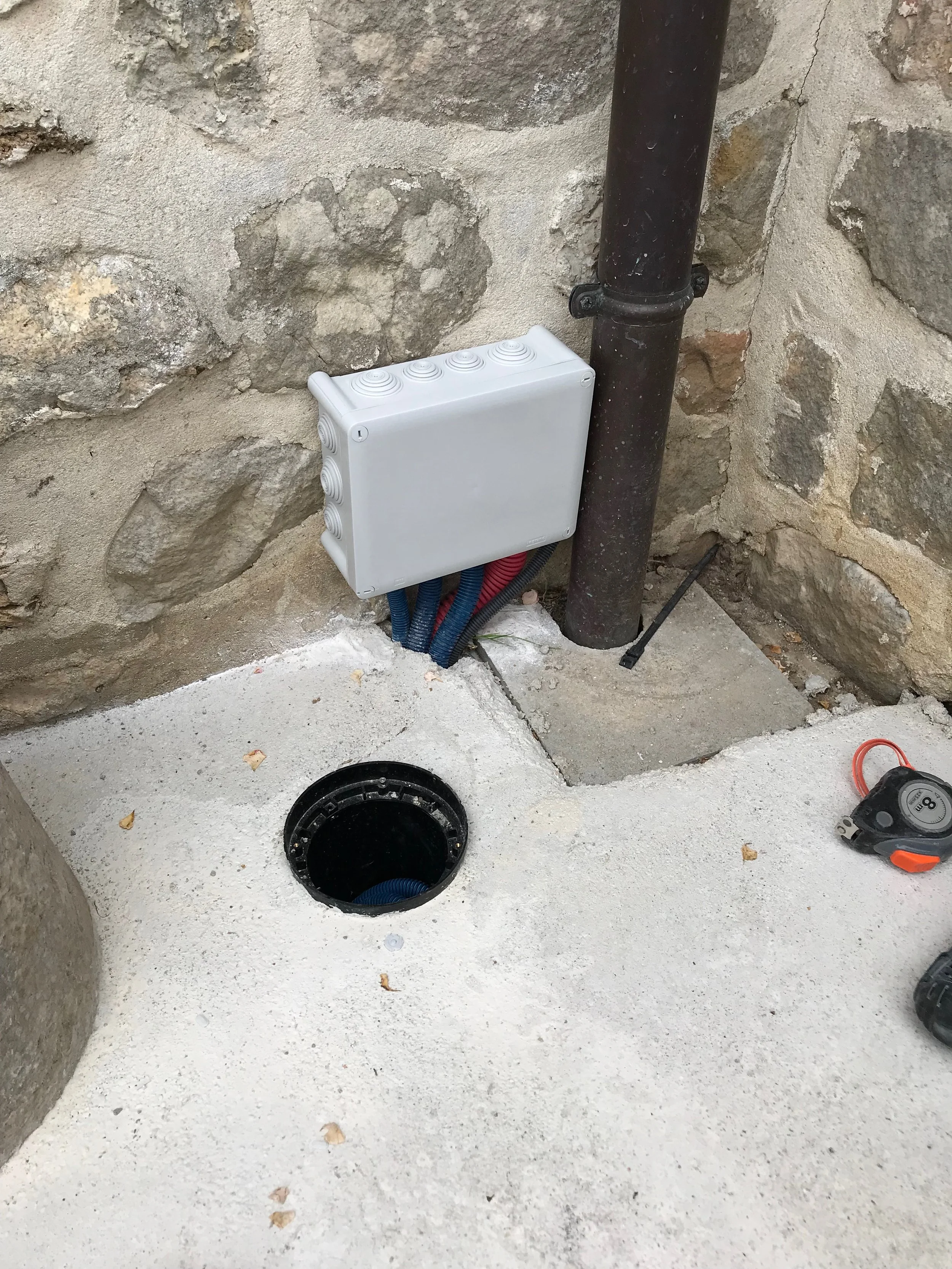 Distribution électrique extérieure avec boîte électrique blanche, tuyaux rouges et bleus, drain ou trou dans le sol, mur en pierre en arrière-plan.