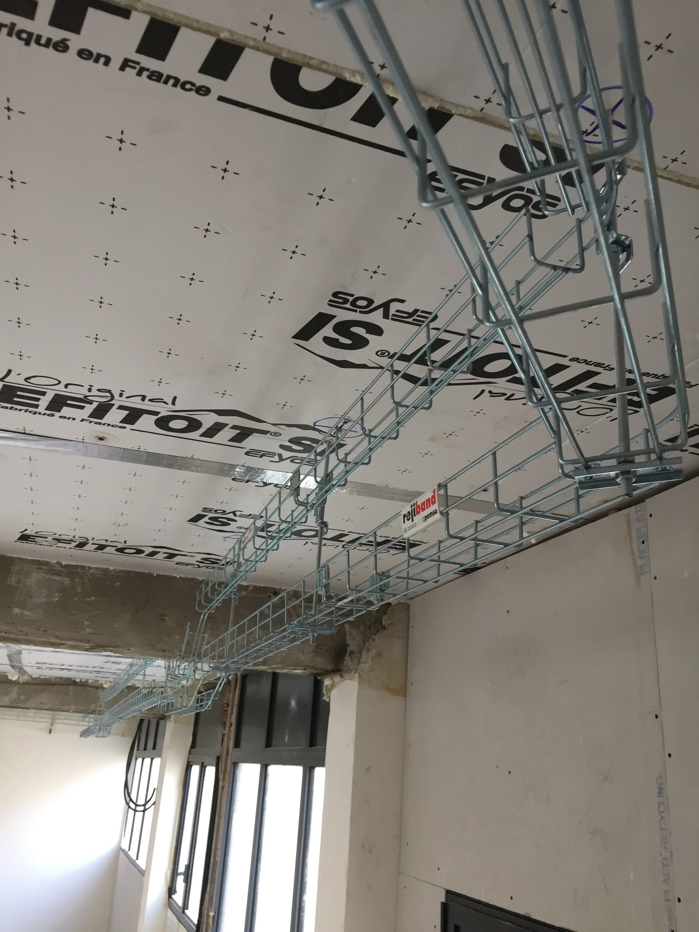 Travée métallique fixée au plafond en construction, avec une isolation et des fenêtres en arrière-plan.