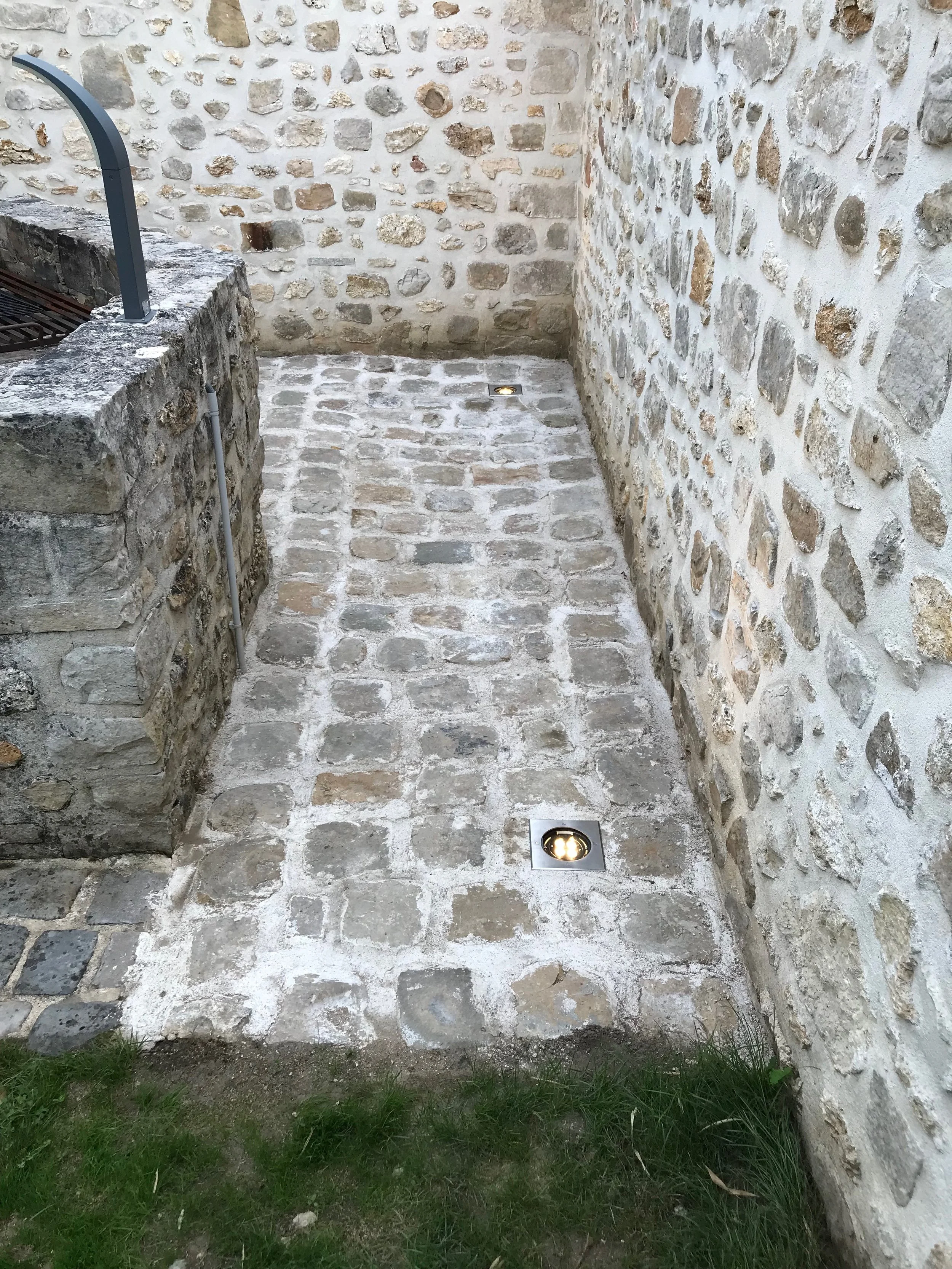 Chemin pavé en pierre avec éclairages encastrés, entouré de murs en pierre. À gauche, une structure en pierre avec un grillage au-dessus.