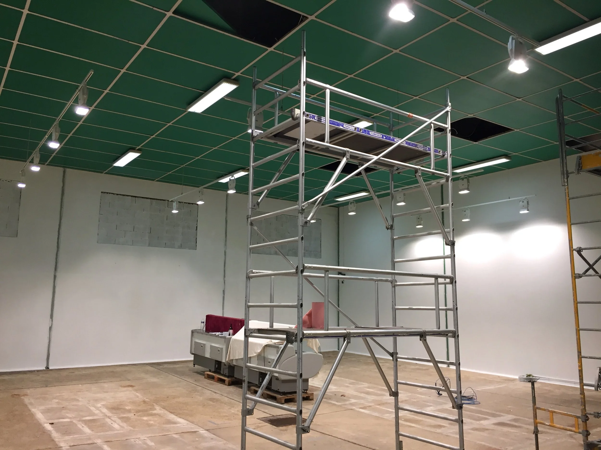Une salle en cours de rénovation avec des échafaudages en métal, un fauteuil médical, et des murs blancs, au plafond un faux plafond avec des lumières suspendues.