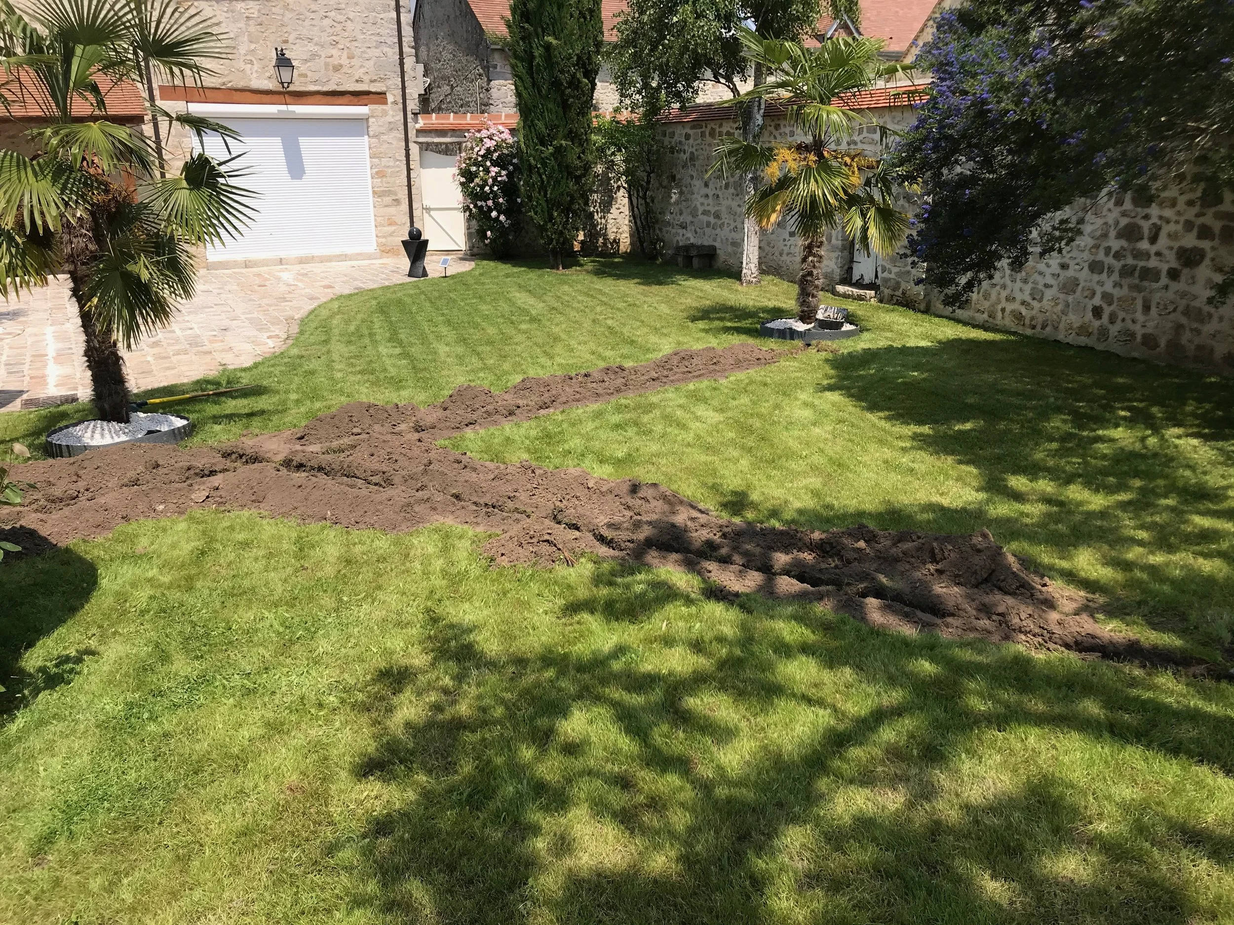 Jardin en train d'être creusé, avec des trous pour planter des arbres ou des plantations, entouré de pelouse et de murs en pierre, avec des arbres et des plantes