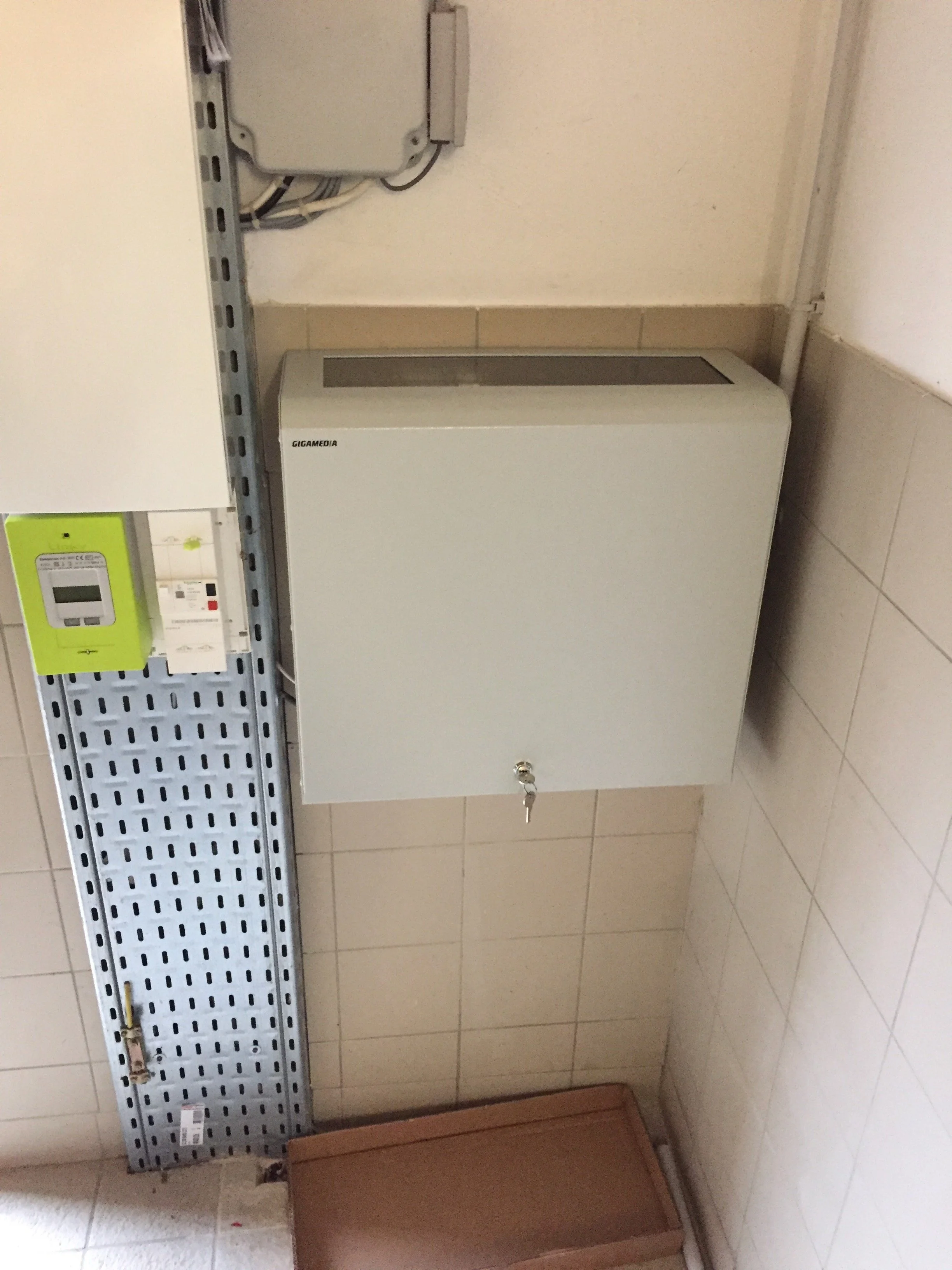 Une armoire métallique grise avec un cadenas, placée au mur dans une pièce avec des carreaux beige au sol et au mur, avec un tableau électrique et un panneau de distribution électrique à gauche.