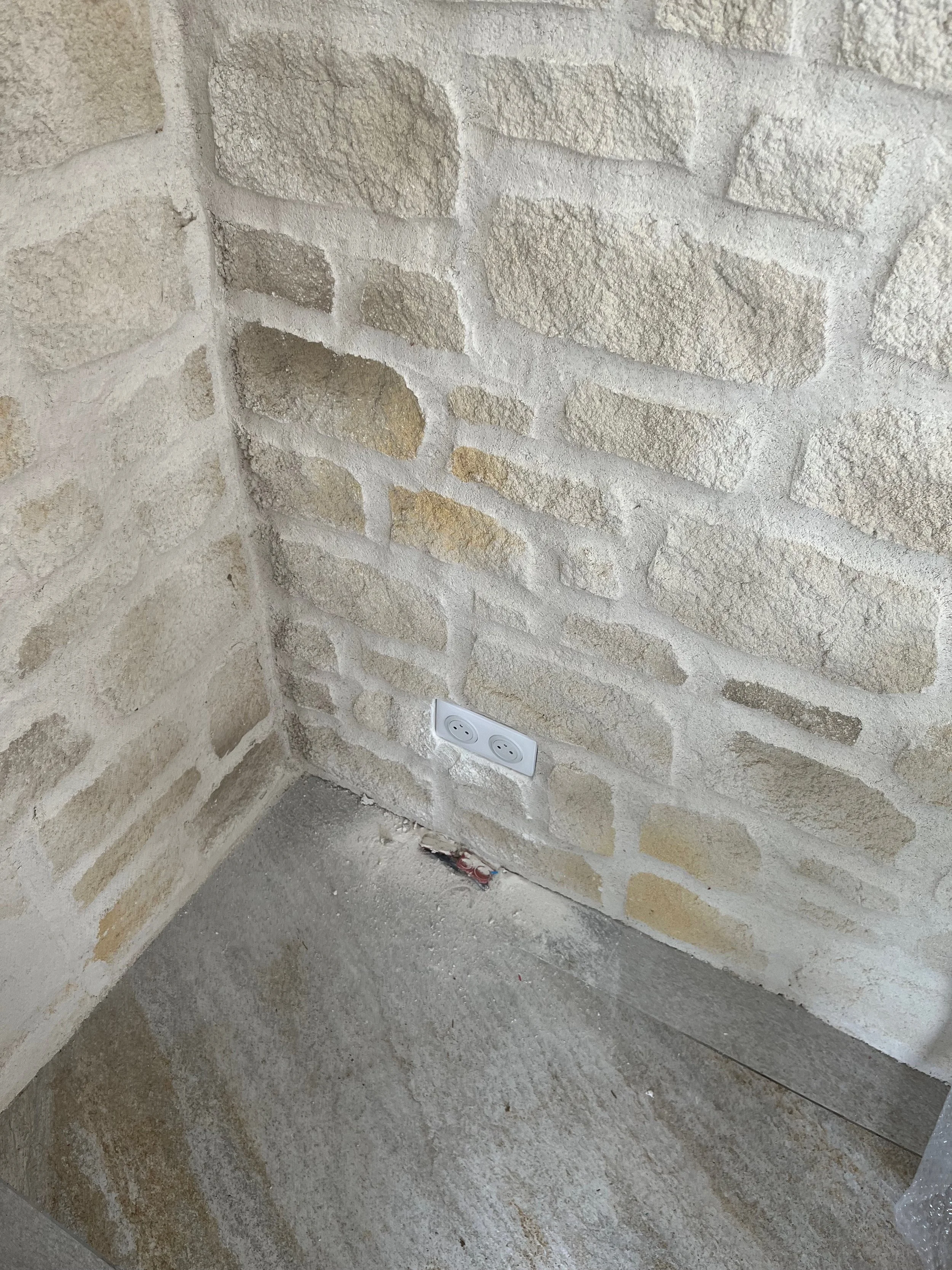 Prise électrique encastrée dans un mur en pierre beige, au coin d'un mur et au sol en béton.
