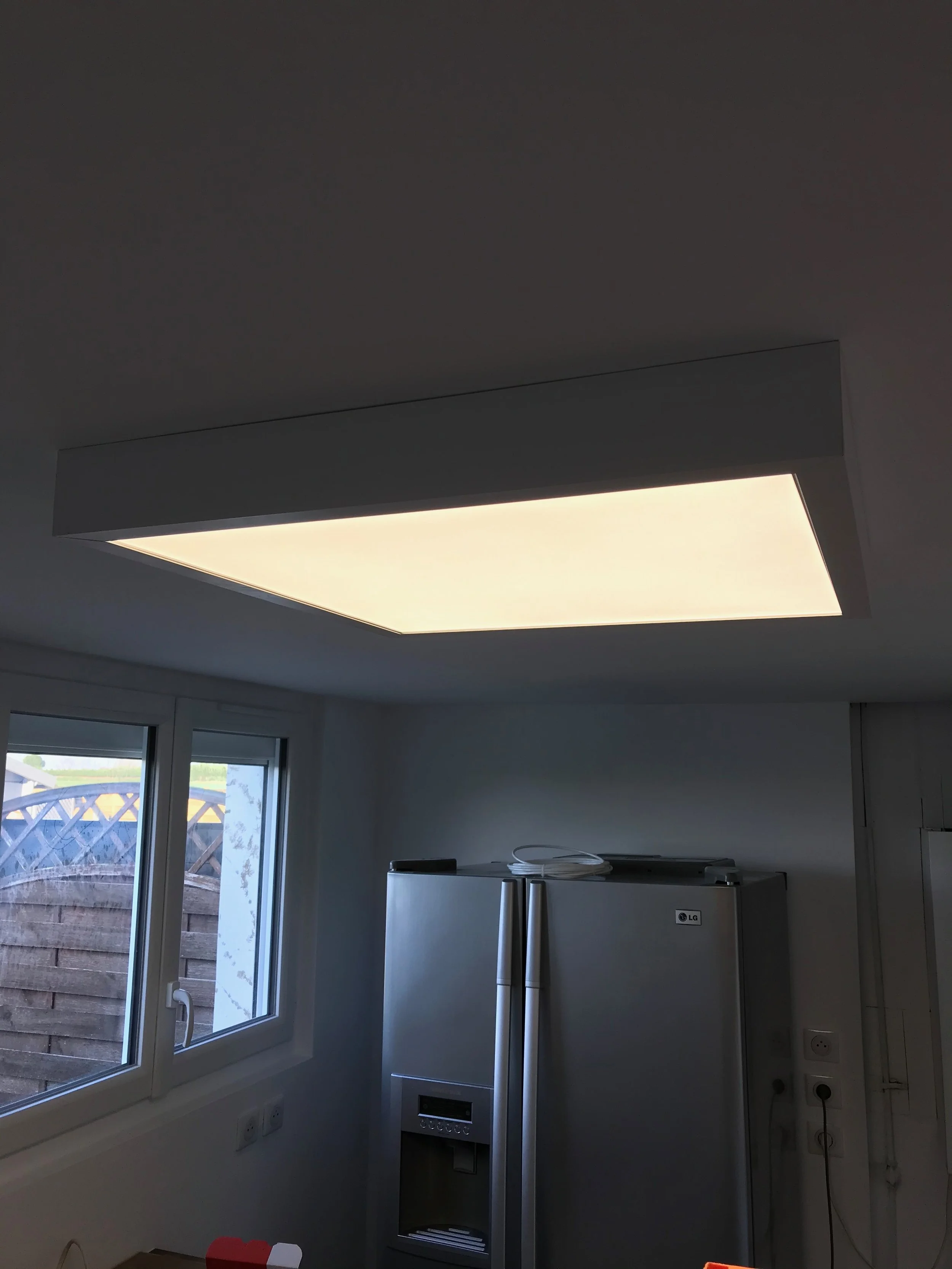 Une vue intérieure d'une cuisine avec un plafond illuminé, une fenêtre laissant passer la lumière naturelle et un réfrigérateur en acier inoxydable.