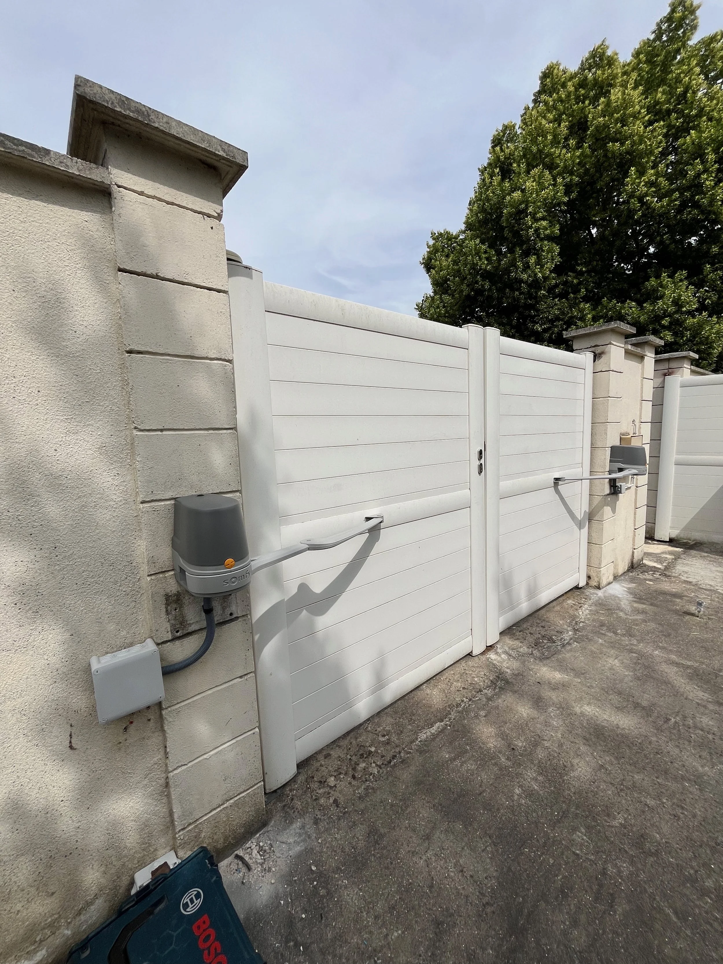 Portail automatique blanc avec deux moteurs côté paroi en béton, dans une cour extérieure, avec un arbre vert en arrière-plan.