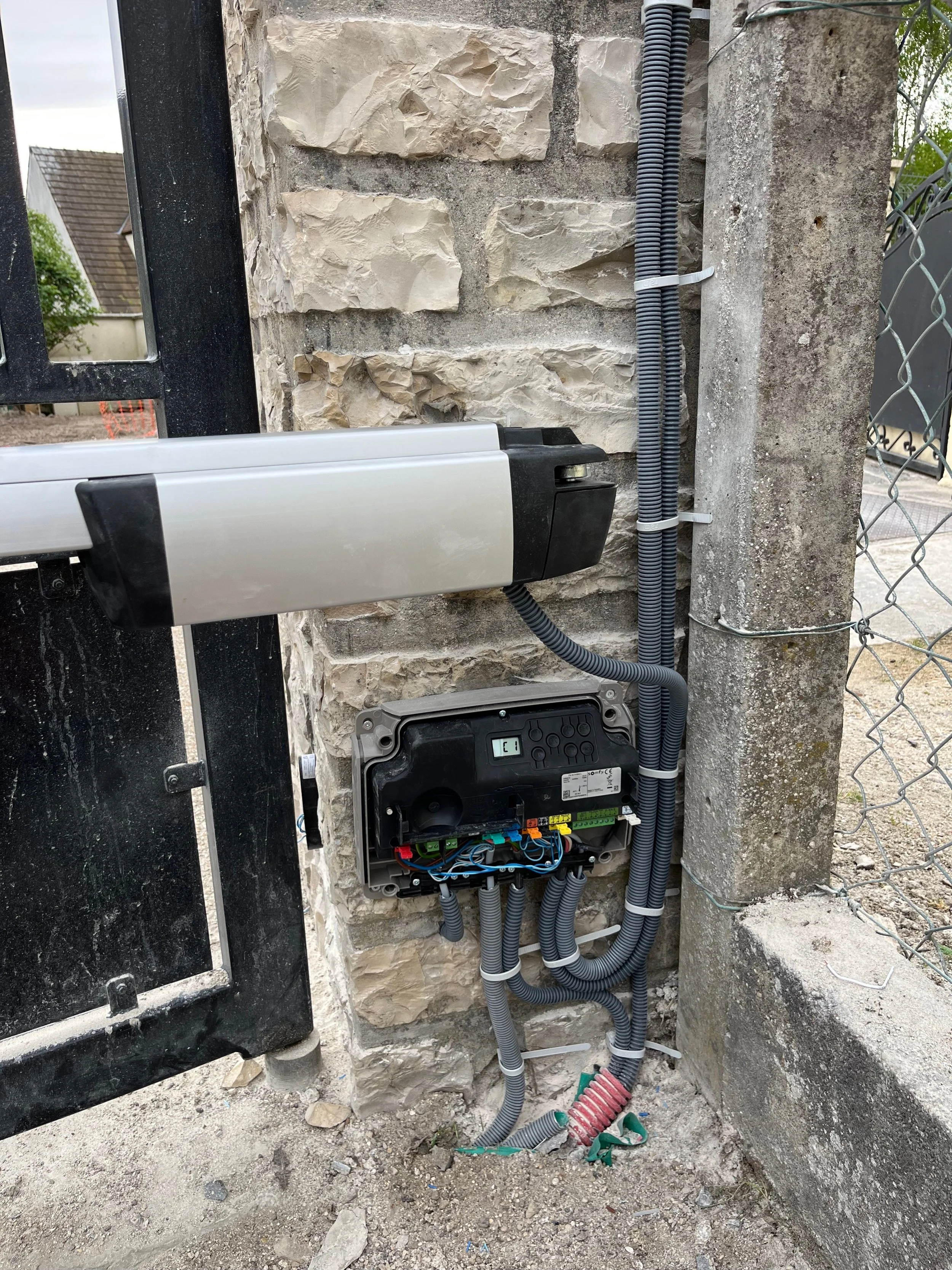 Installation électrique extérieure avec un boîtier de commande, des câbles protégés par des conduits, fixés à un mur en pierre et un poteau en béton.
