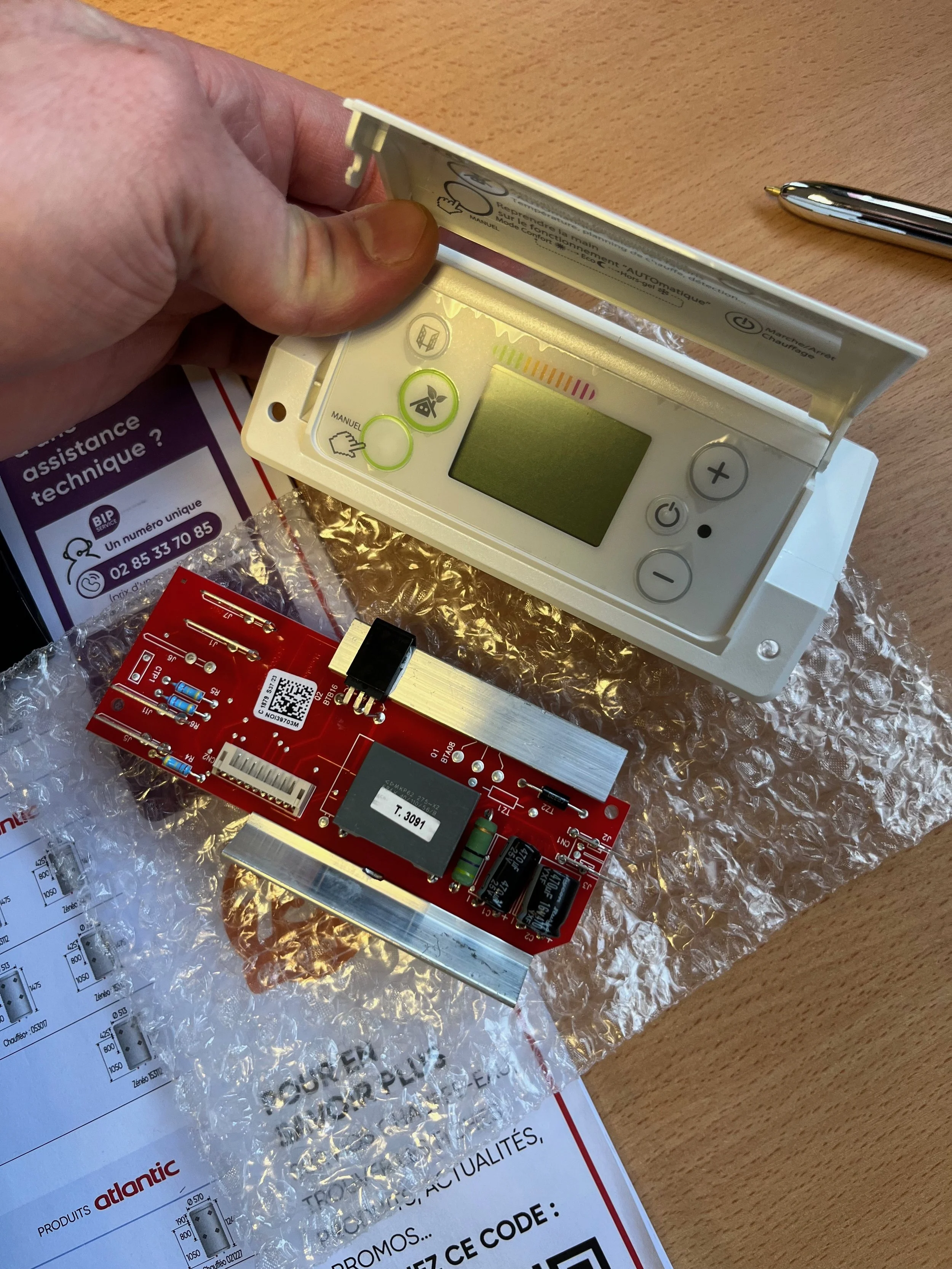 Gros plan sur un module électronique rouge avec composants électroniques, et un appareil de mesure avec un écran digital blanc, boutons + et - et autres contrôles, placé sur un papier avec un diagramme. La main d'une personne tient une partie de l'ap