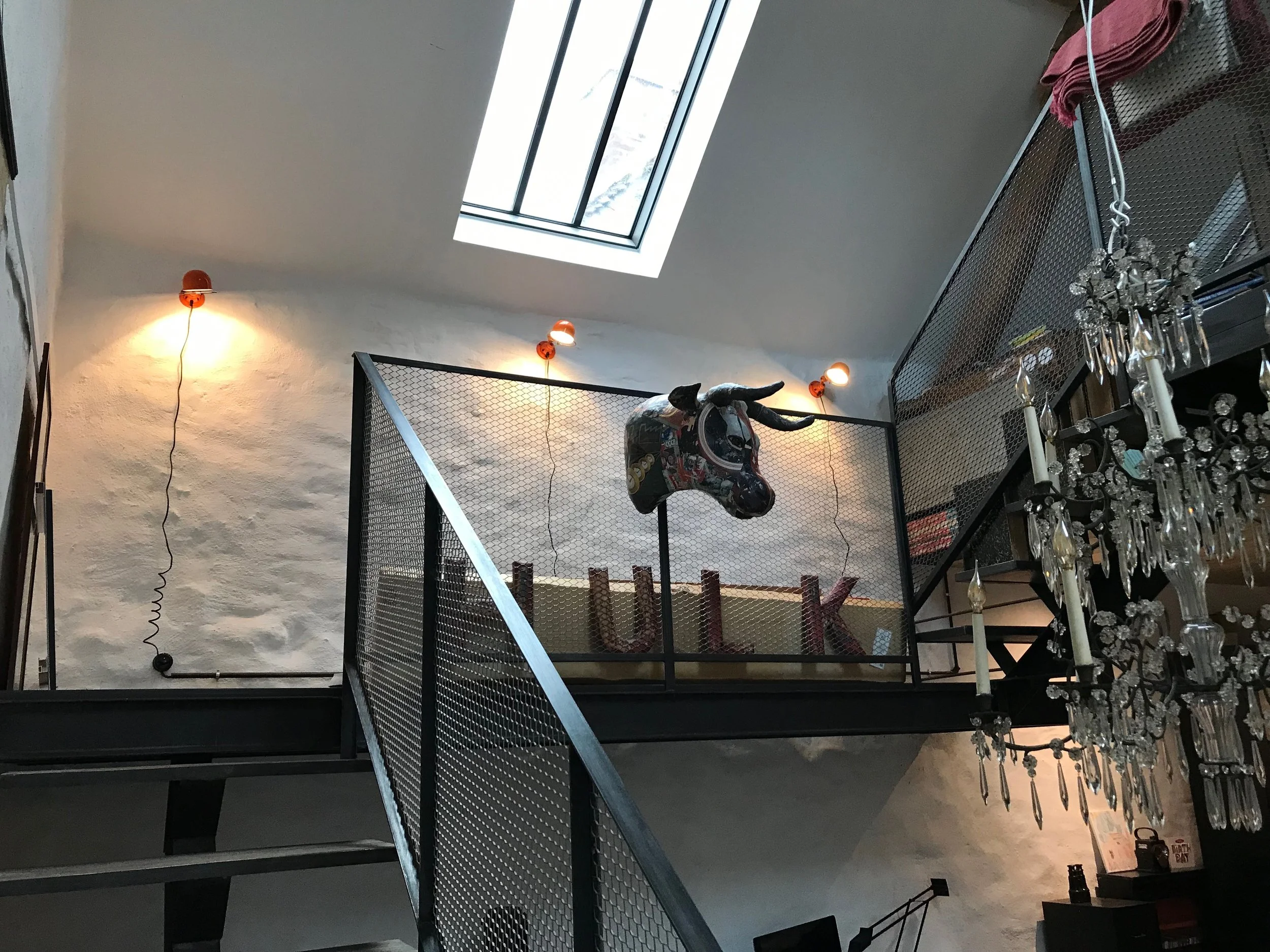 Intérieur d'un loft avec un mur blanc, un escalier en métal noir, un mur en pierre, un puits de lumière dans le plafond, une tête de taureau en décoration suspendue et un chandelier en cristal.